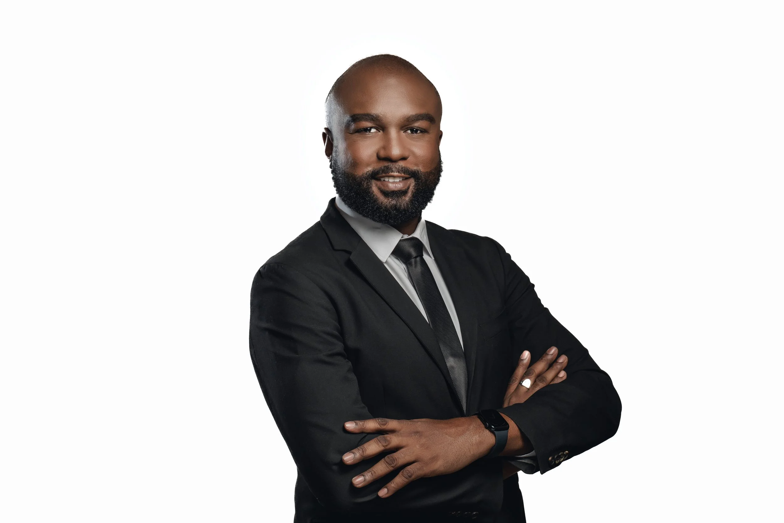 A man with a dark beard and a shaved head wearing a black suit with a white shirt and black tie, standing with arms crossed against a plain white background.