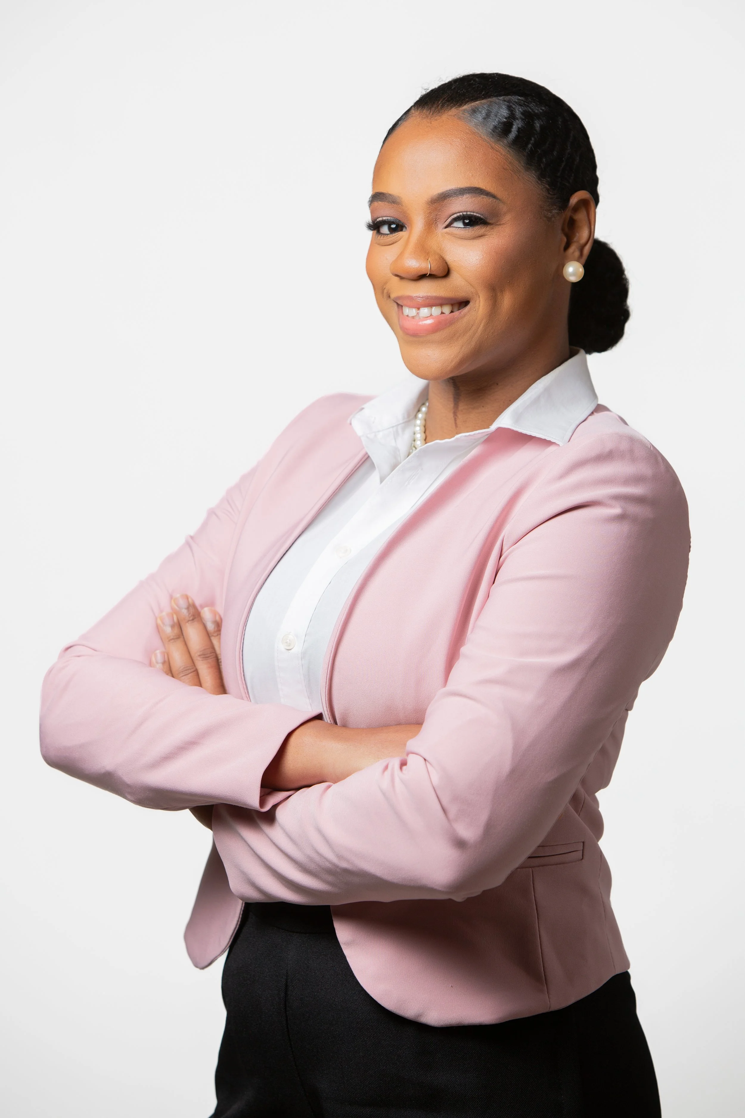 A confident woman in a pink blazer, white shirt, and black skirt, smiling with arms crossed against a plain white background.
