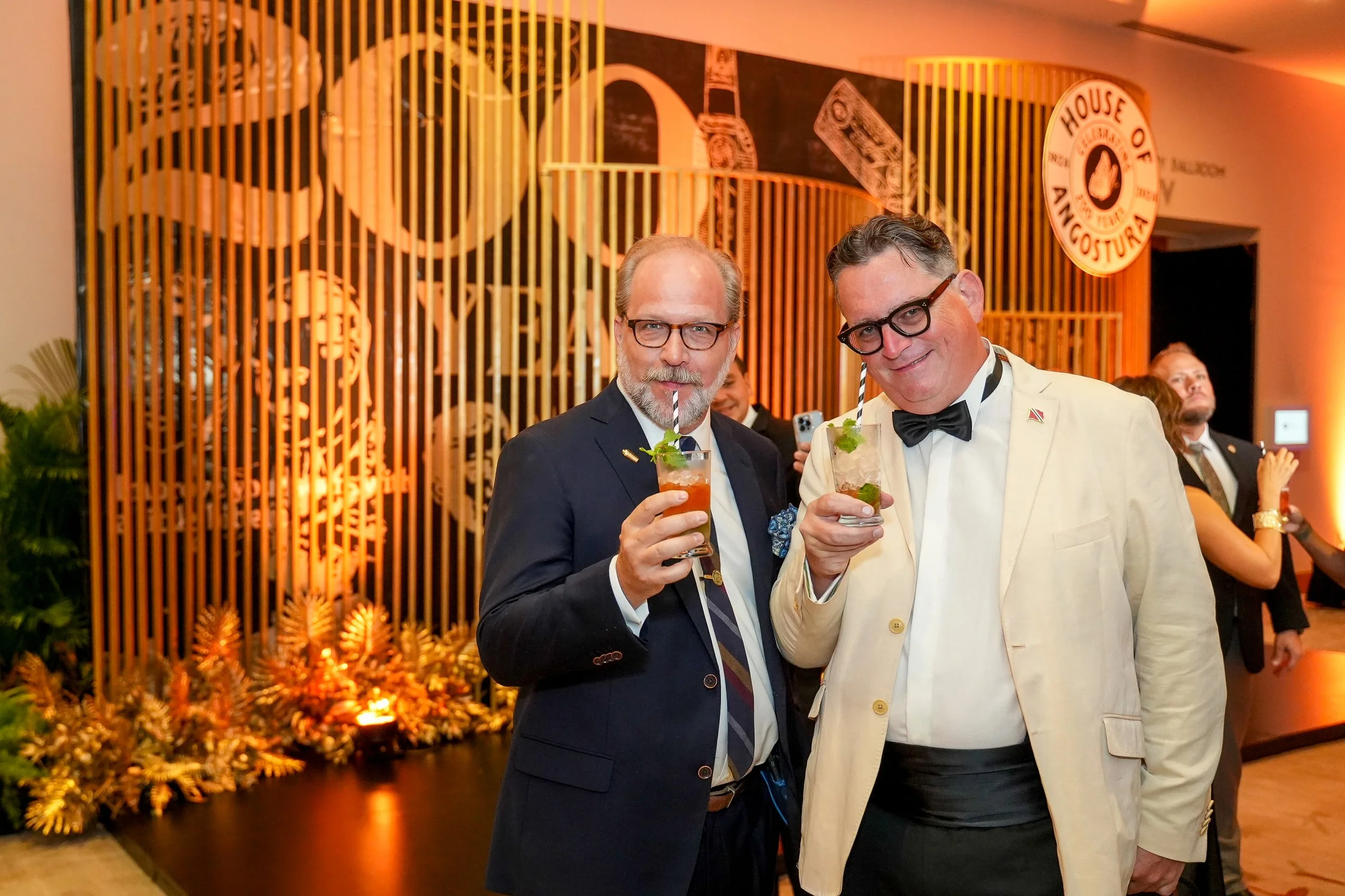 Two men at a formal event holding drinks with striped straws, both wearing glasses and dressed in suits. One man is in a dark suit with a tie, and the other in a light suit with a bow tie. Behind them is a decorative wall with a logo that reads 'Hous