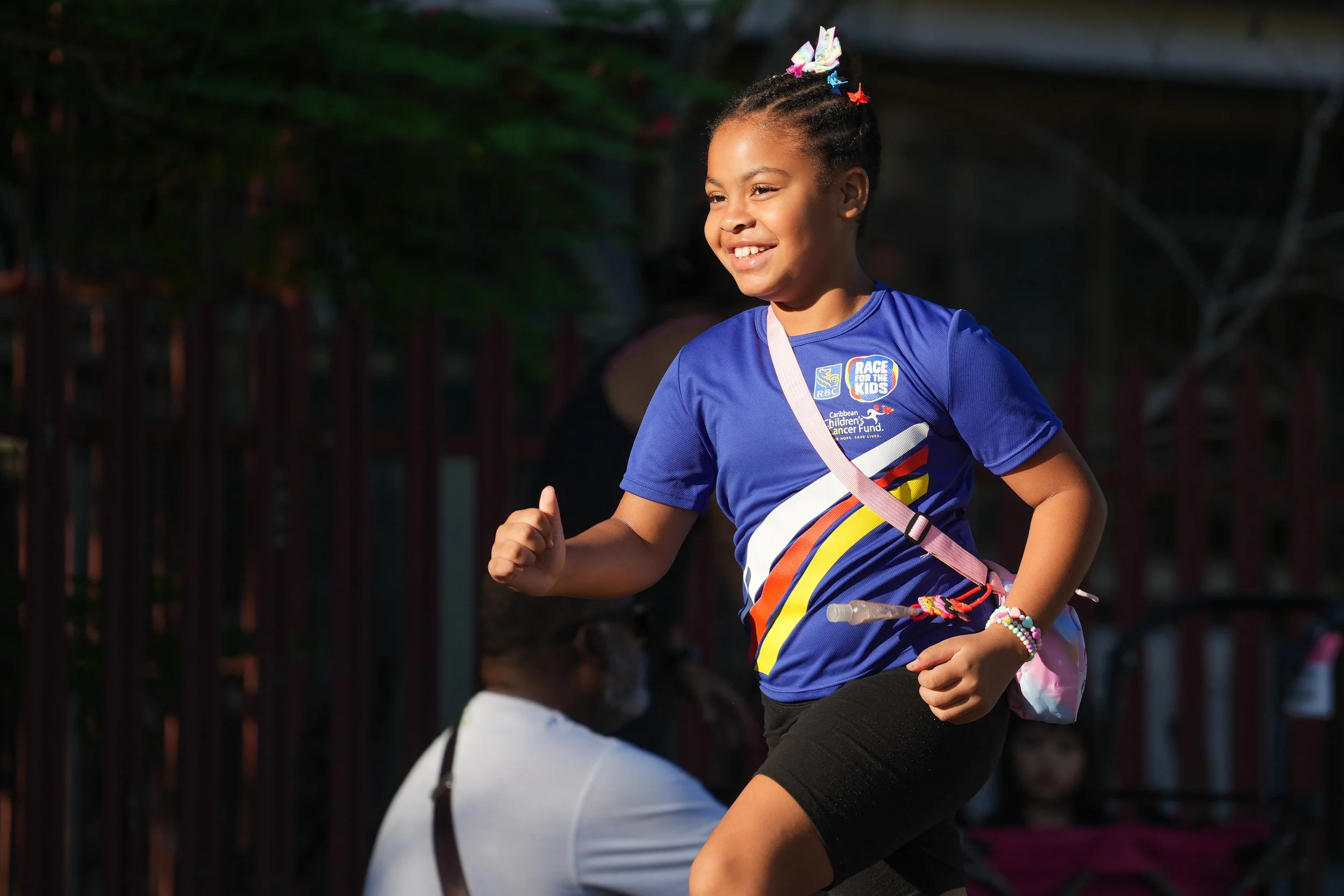 Young girl with braided hair smiling and running outdoors, wearing a blue t-shirt with colorful stripes and a lanyard, participating in a children's race event.