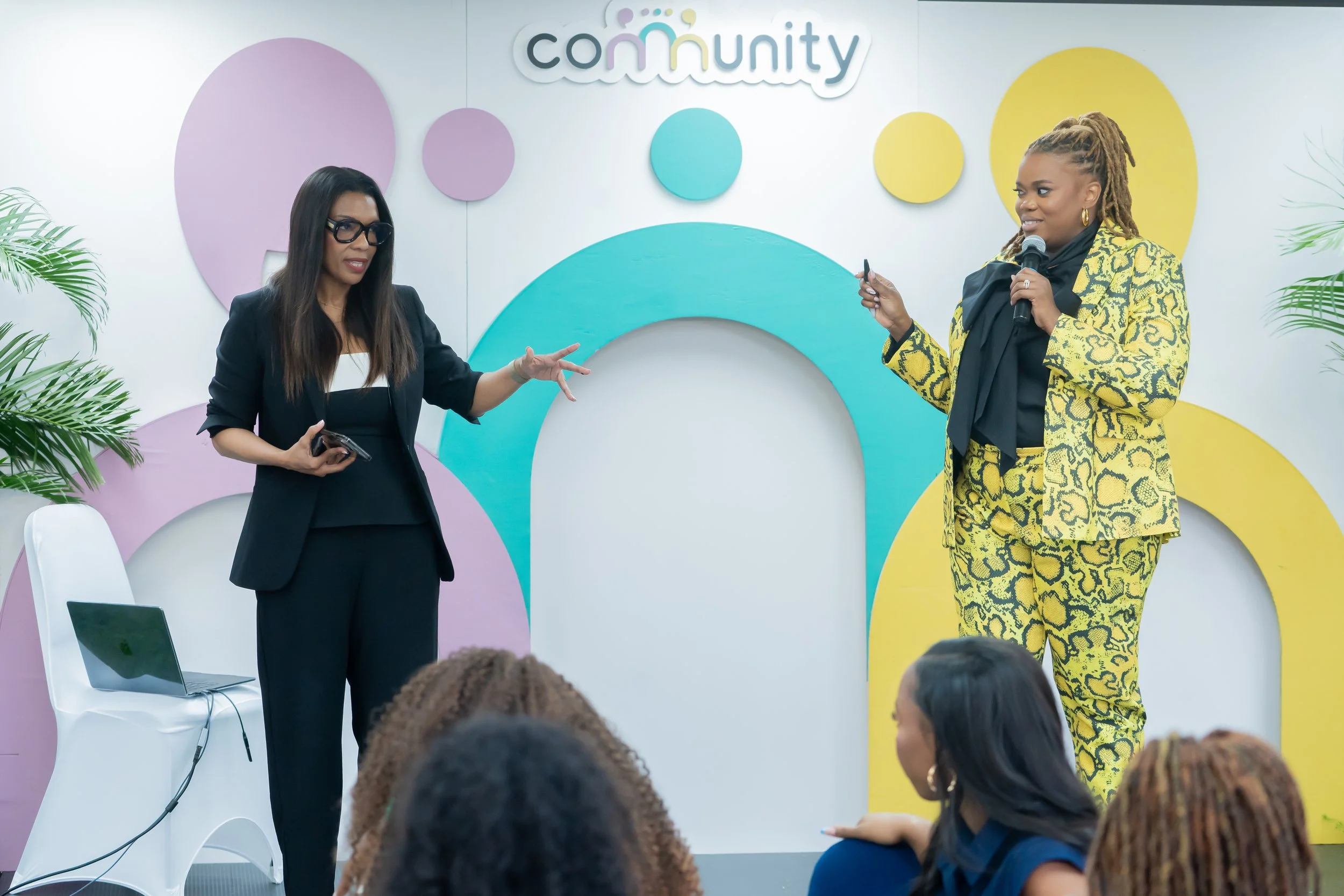 Two women speaking at a community event with an audience. The woman on the left is wearing black glasses, a black blazer, and holding a phone. The woman on the right has braided hair, gold hoop earrings, and is dressed in a yellow and black patterned