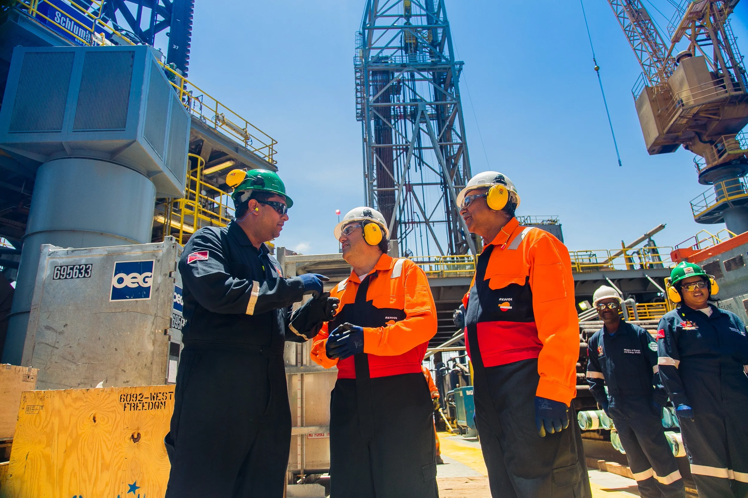 Construction workers in safety gear, such as helmets and ear protection, standing on a construction site with equipment and structures, engaging in conversation.