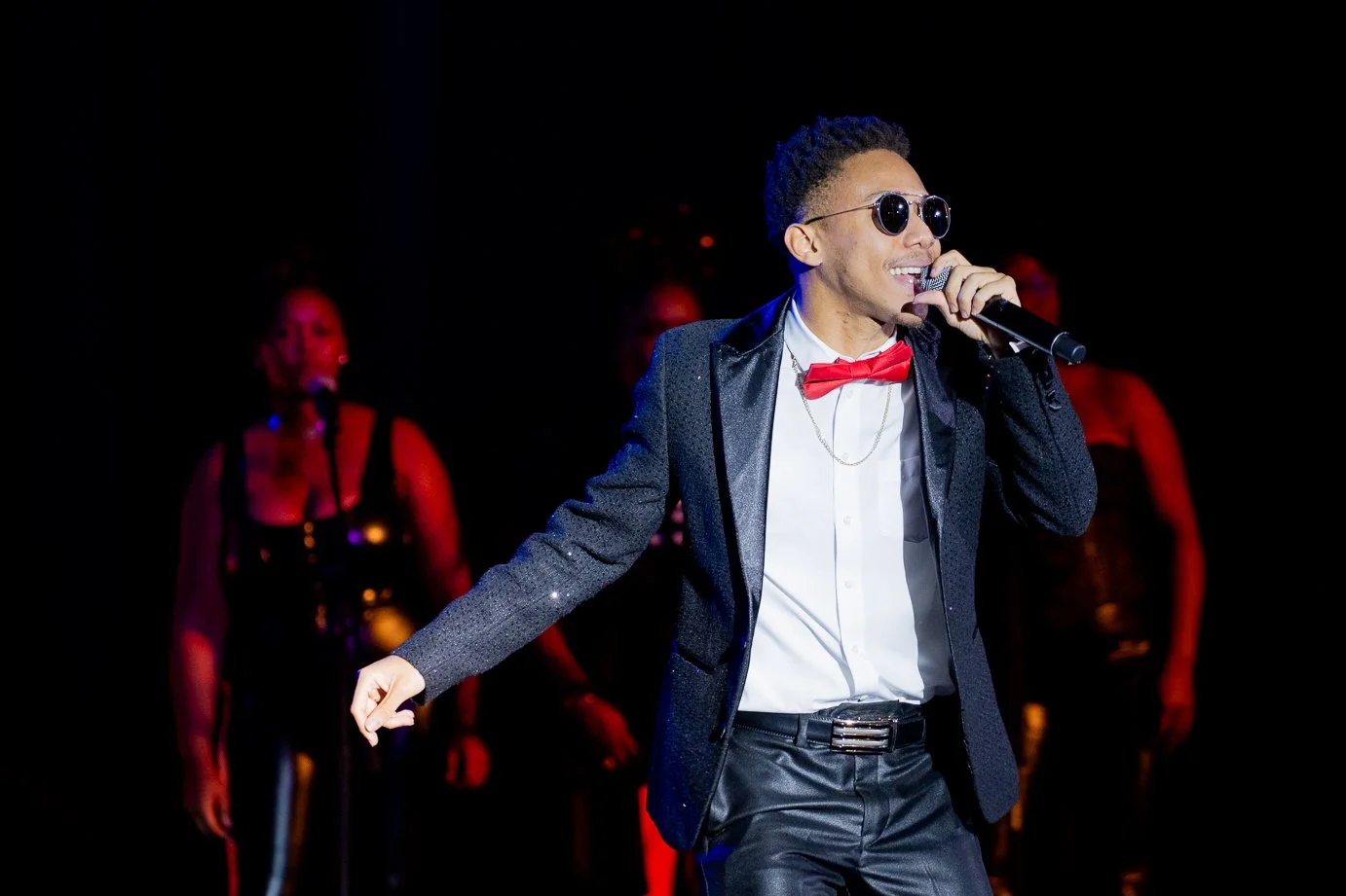 A male singer dressed in a tuxedo with a red bow tie and sunglasses, singing into a microphone on stage, with backup singers in the background.