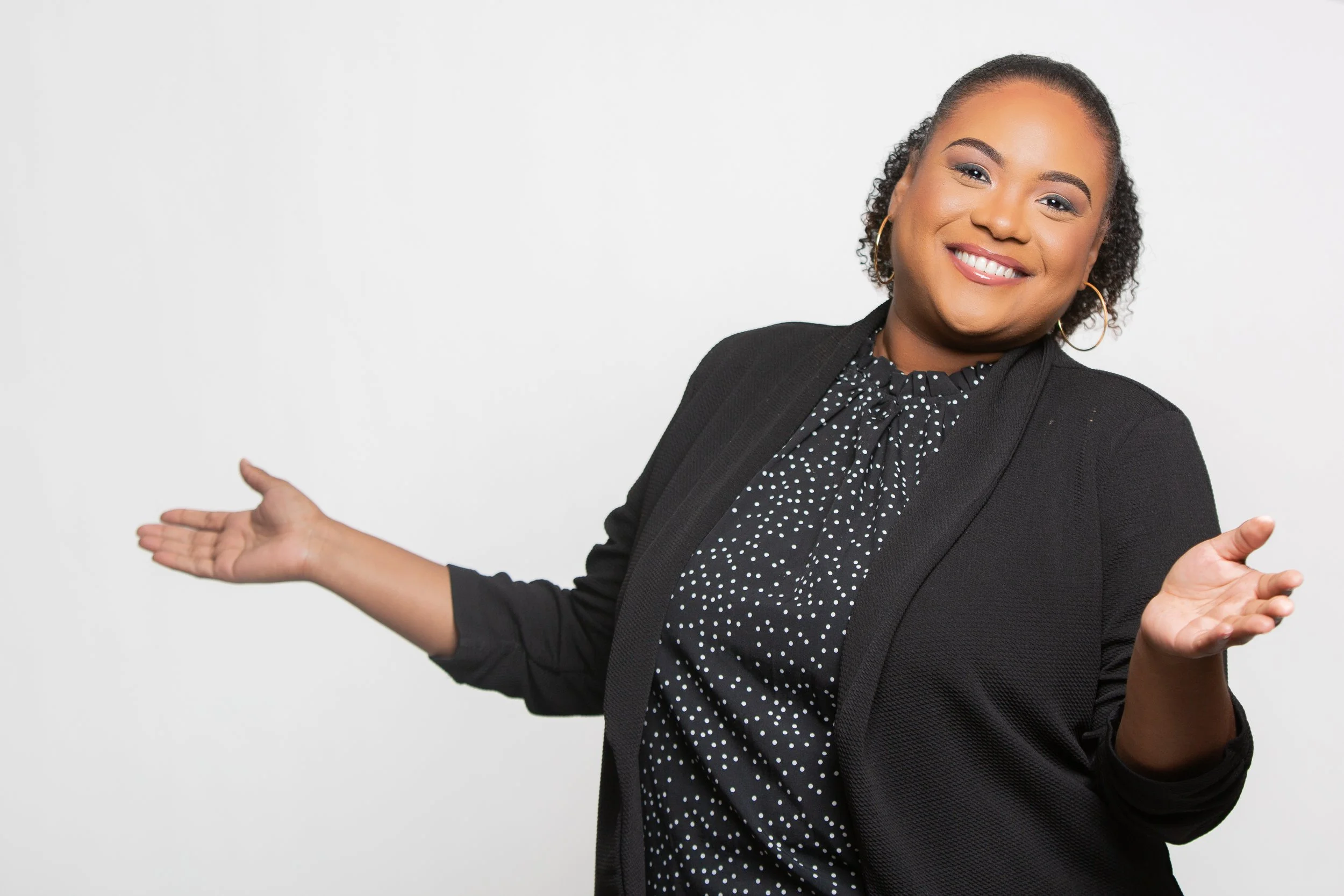 A woman with short curly hair, wearing a dark polka dot blouse and black blazer, smiling and gesturing with open hands against a plain white background.