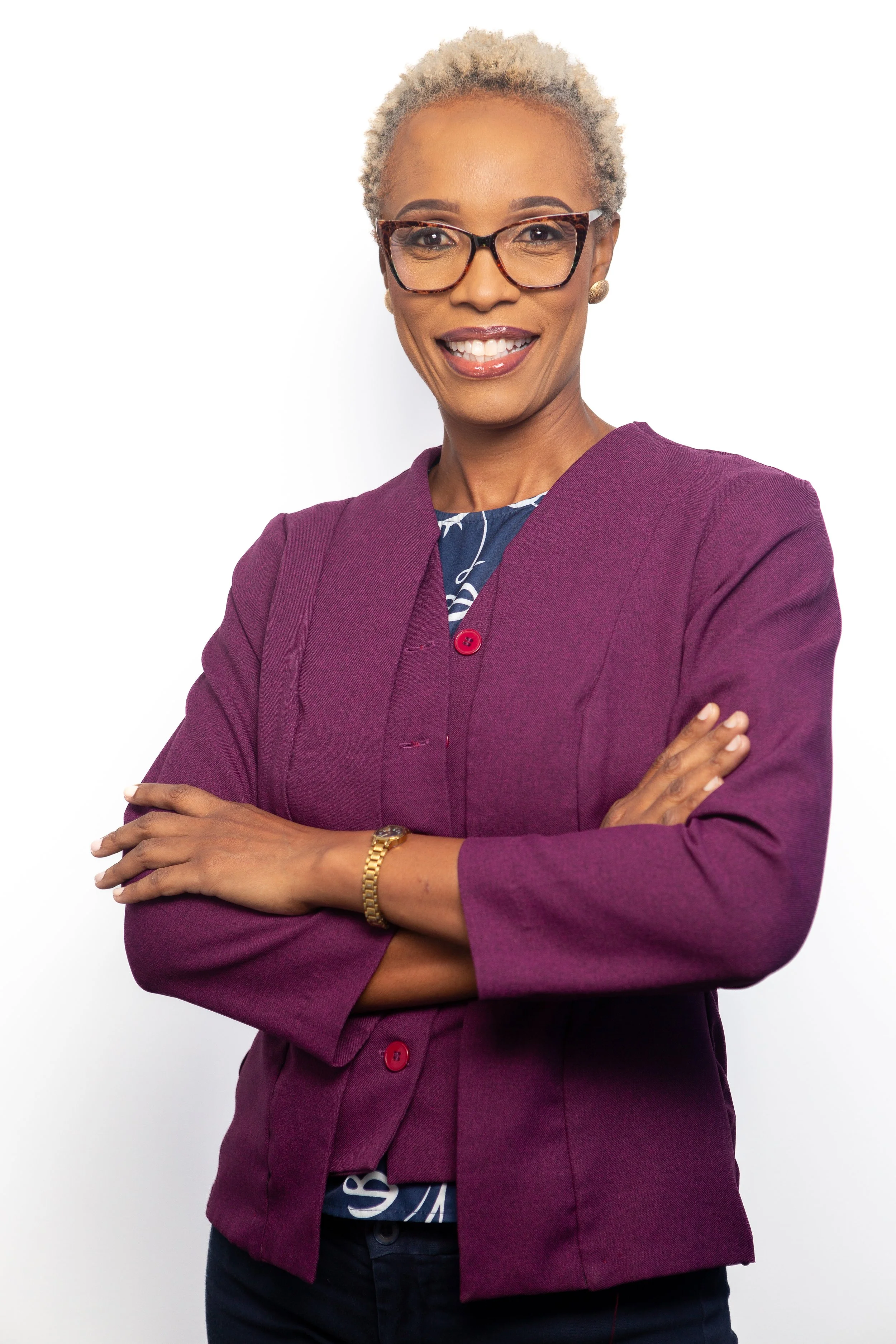 A confident African-American woman with short blonde hair, wearing glasses, a purple blazer, and a patterned top, standing with her arms crossed against a plain white background.