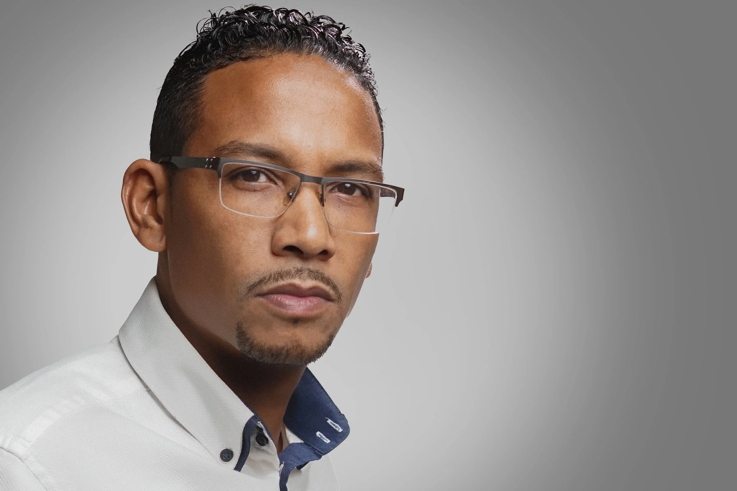 Close-up of a man wearing glasses and a white hat with a dark blue inner lining, dressed in a white blazer and blue shirt, against a gray background.