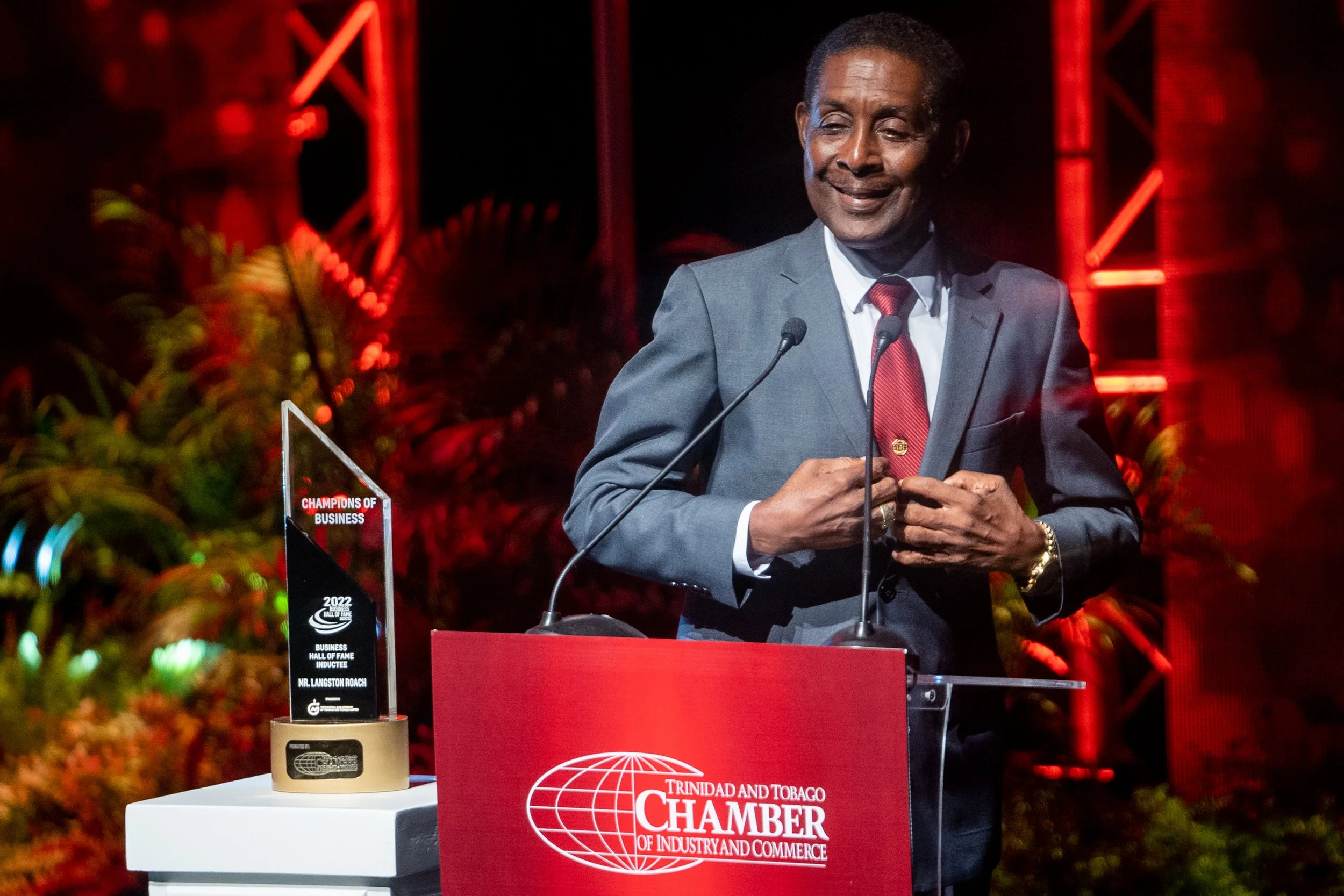 Man in a suit standing at a podium with the Trinidad and Tobago Chamber of Industry and Commerce logo, smiling, with a trophy on a table beside him, at an awards event.