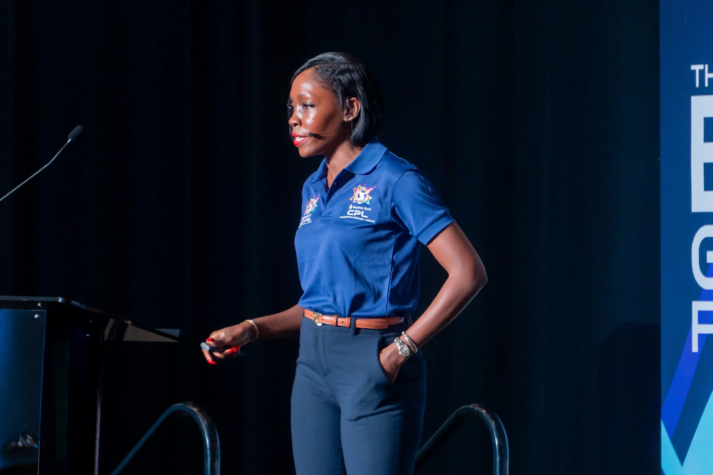 A woman with short black hair wearing a blue polo shirt with logos, standing on stage at a presentation or conference, with a microphone headset, and holding a remote control, in front of a dark background.