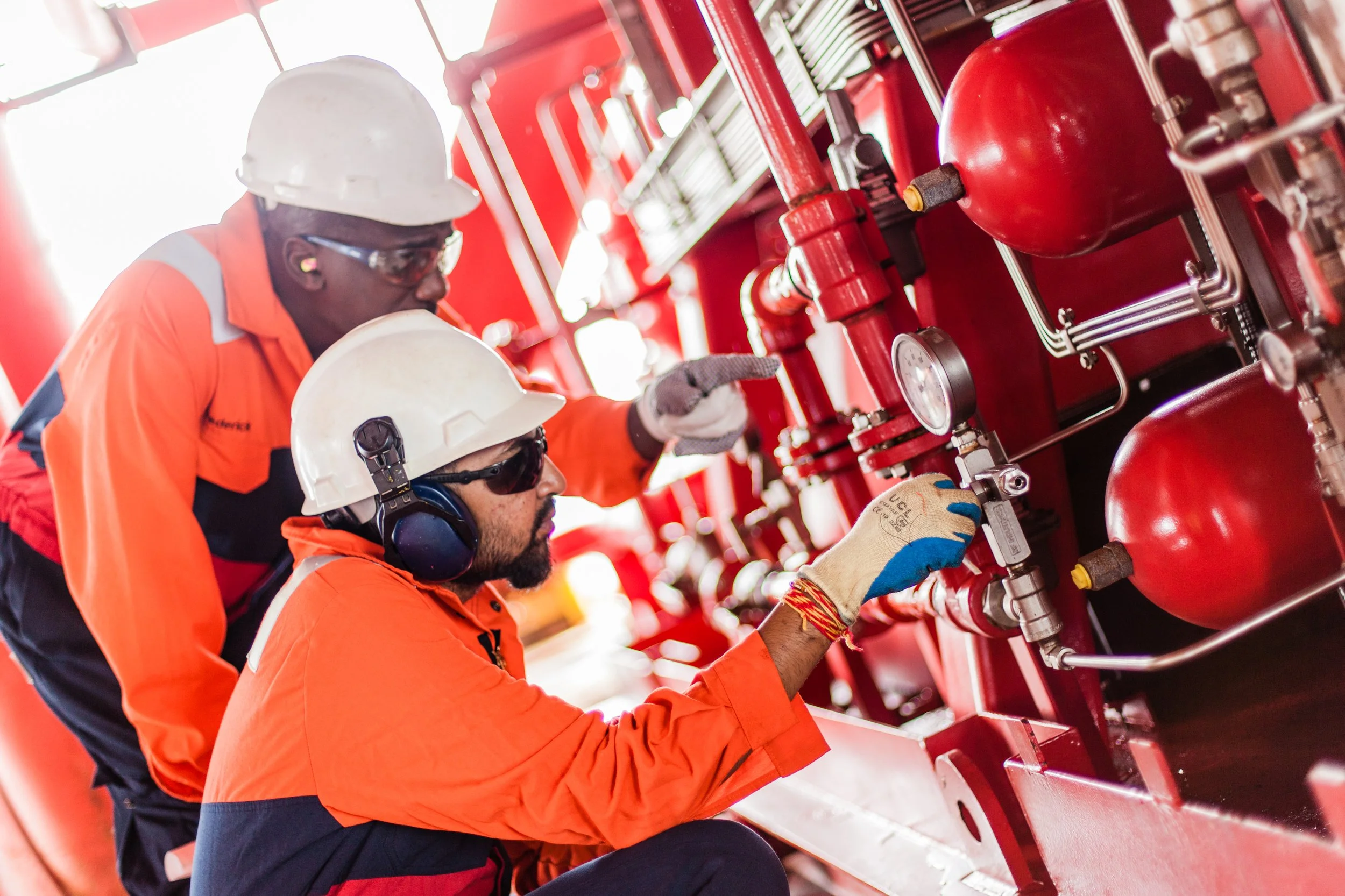 Two workers wearing orange safety uniforms, hard hats, and safety glasses inspecting red fire suppression system pipes in an industrial setting.