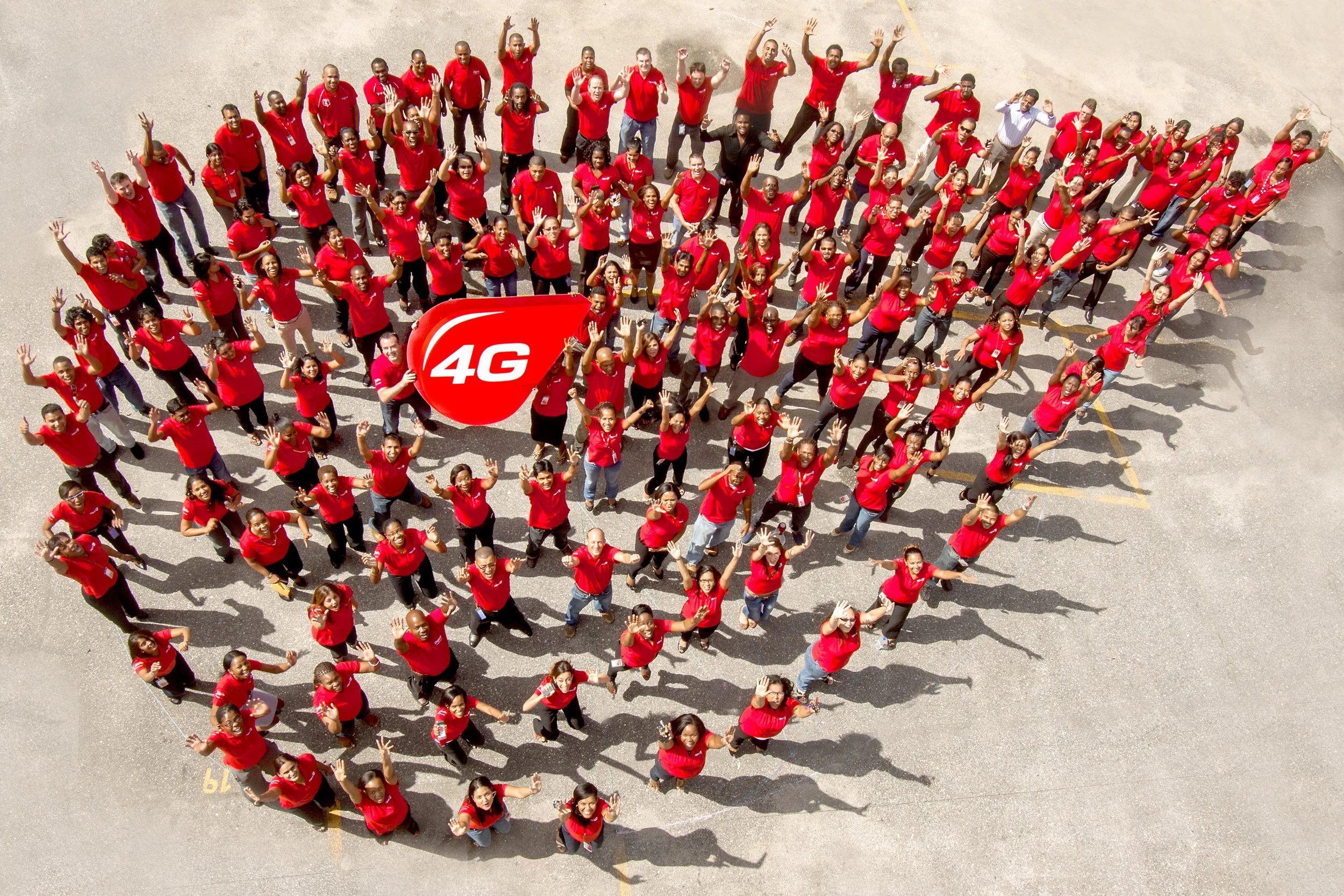 A large group of people wearing red shirts, standing together outdoors on a paved area, forming a heart shape around a red 4G sign, and waving at the camera.
