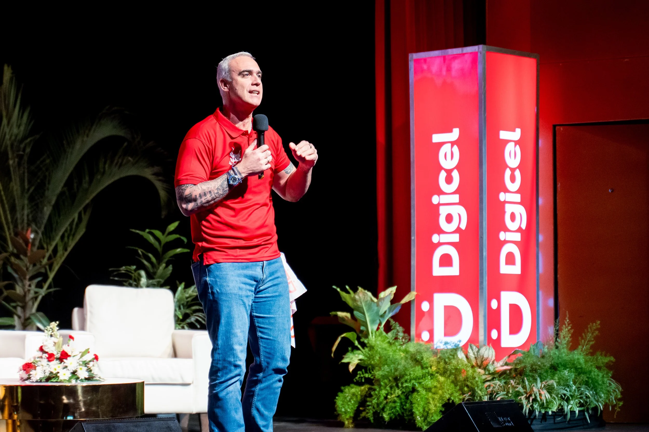 A man with tattoos on his arms wearing a red shirt speaking into a microphone on stage with red banners and plants in the background.
