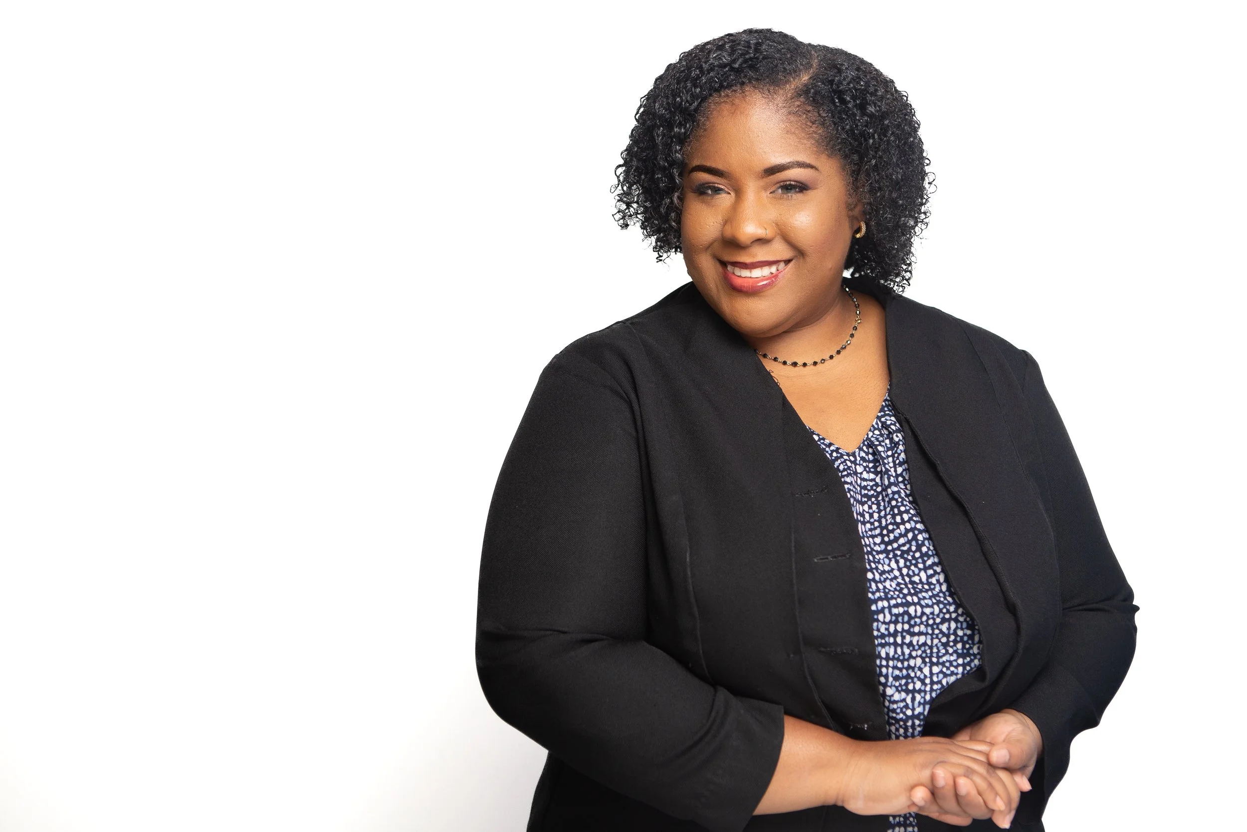 An African American woman with curly hair wearing a black blazer and patterned blouse, smiling with arms crossed against a white background.
