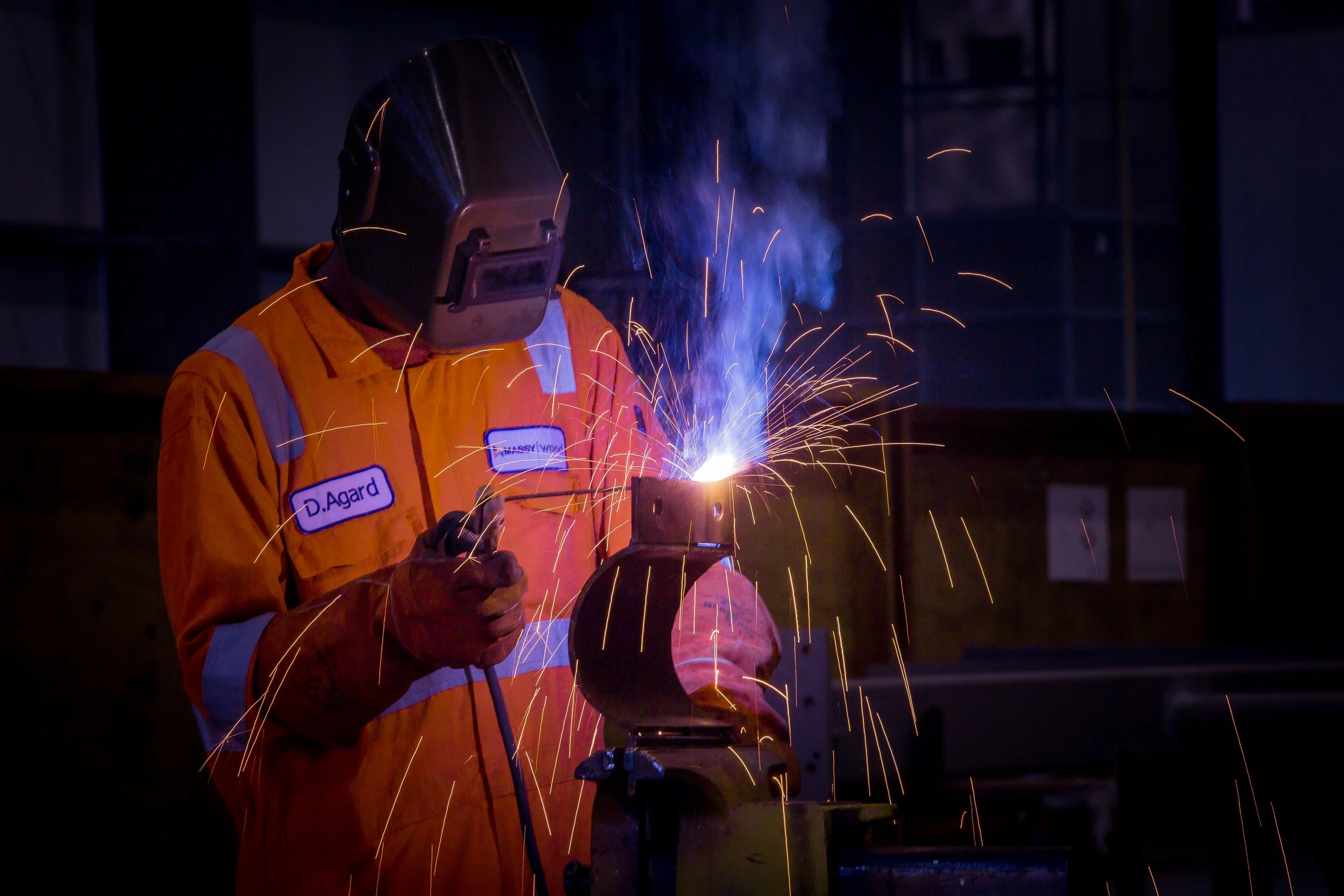 A worker wearing a welding helmet and orange safety jacket is welding metal, creating sparks and blue smoke in an industrial setting.
