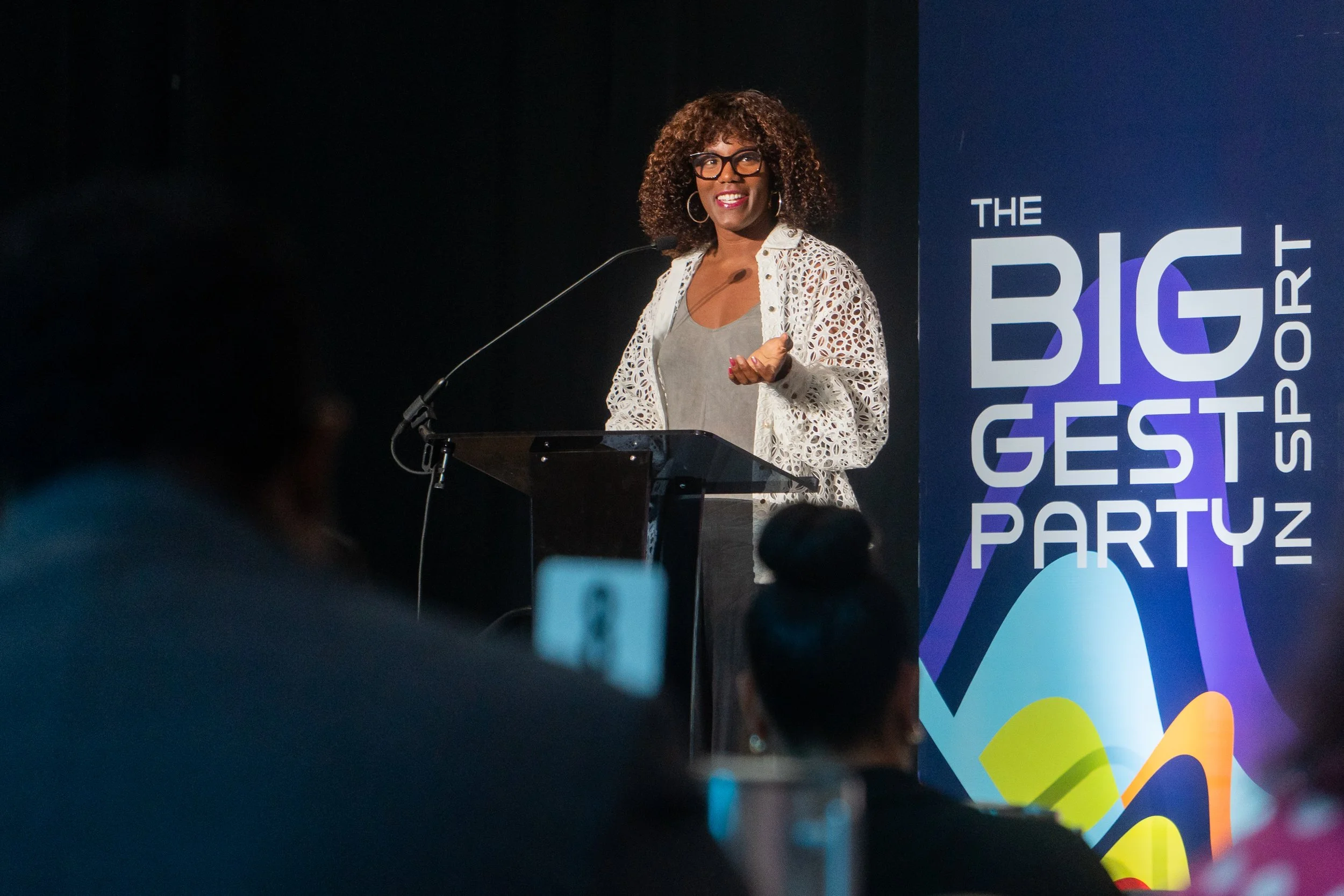A woman with curly hair and glasses smiling and speaking at a podium during 'The Biggest Party' event, with a colorful geometric background.