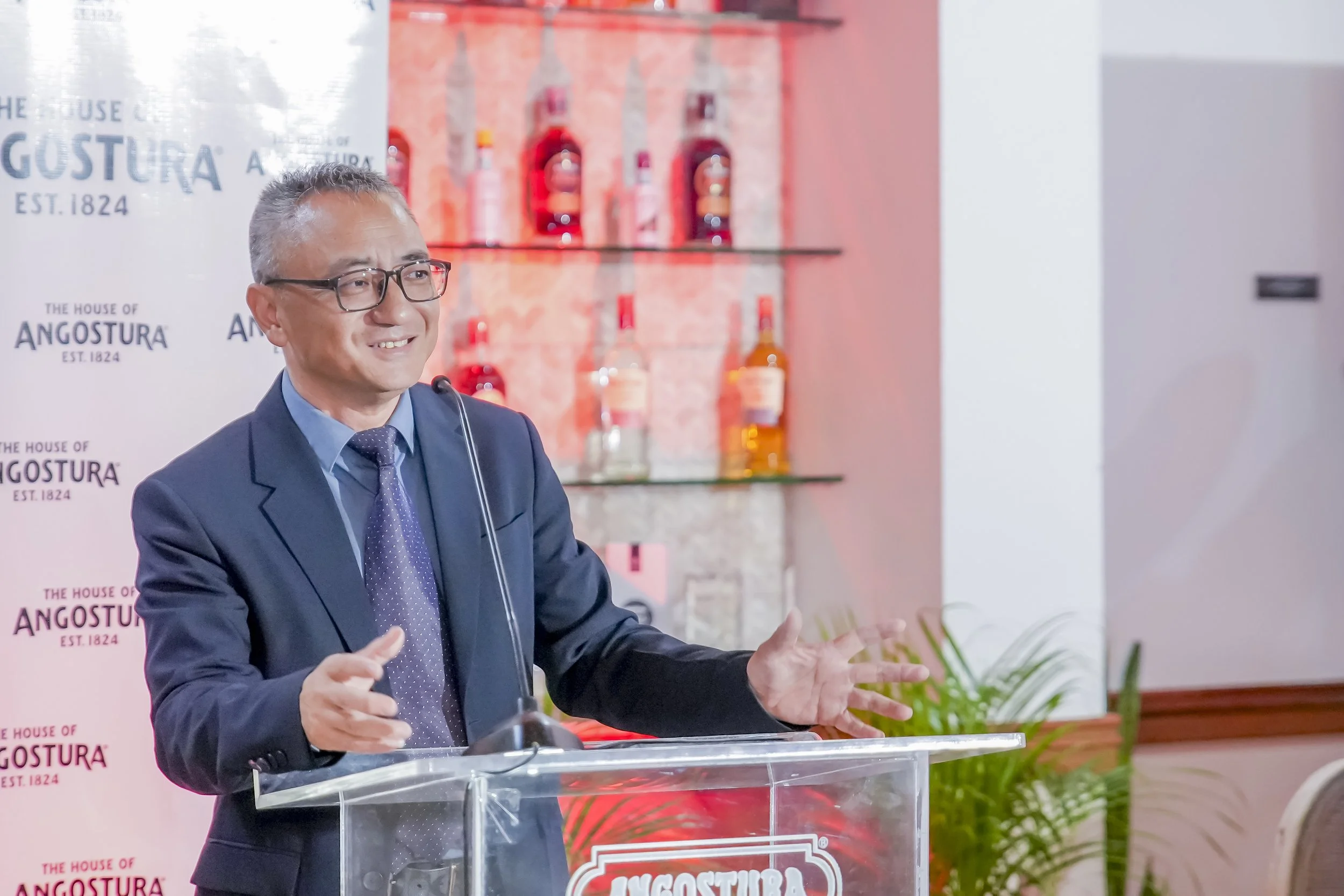 A man in a suit and glasses speaking at a podium during a presentation with a background displaying bottles and a banner with the text 'The House of Angostura, Est. 1824'.