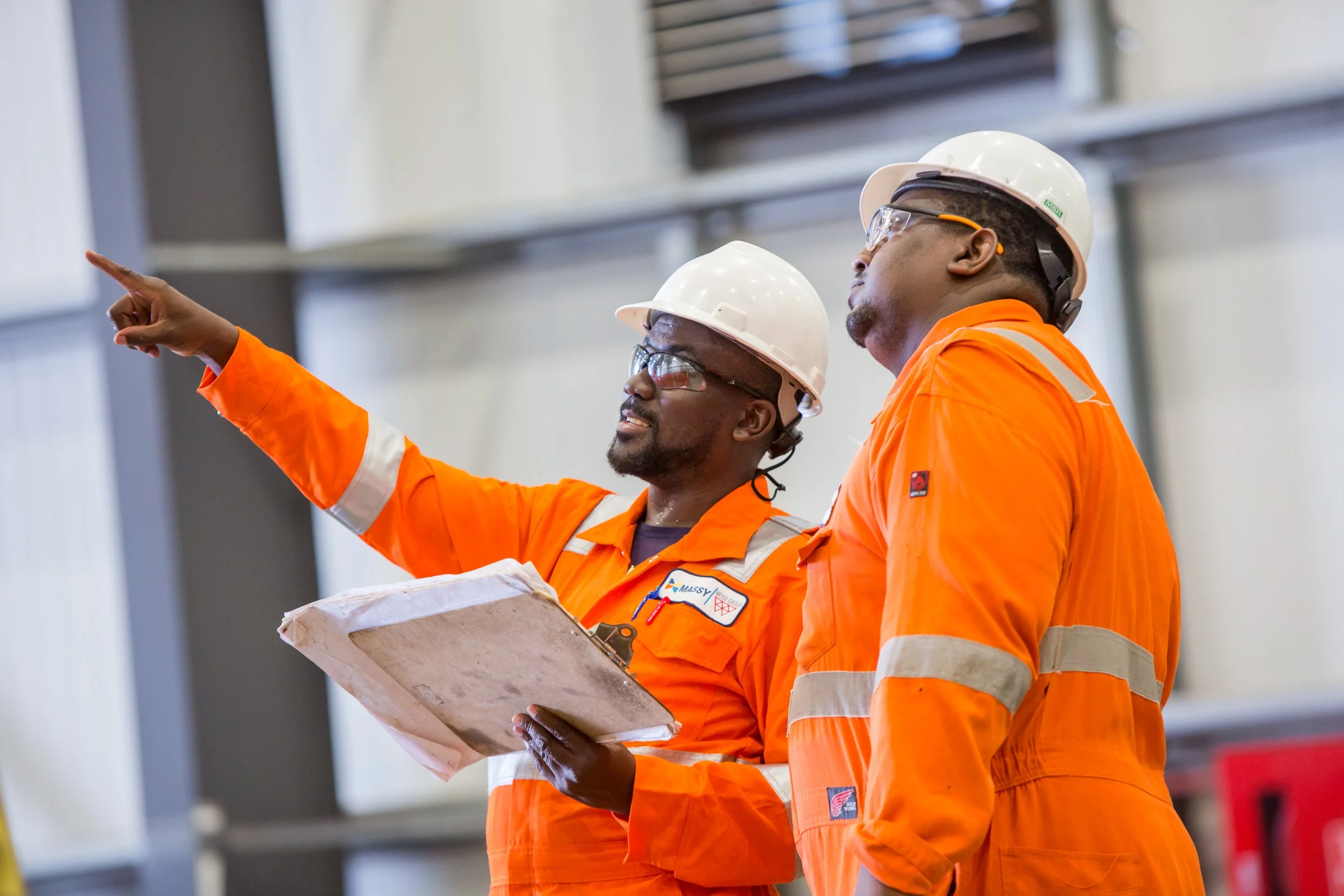 Two construction workers wearing orange safety uniforms and white hard hats are looking at something, with one pointing to the side while the other holds a clipboard or document.