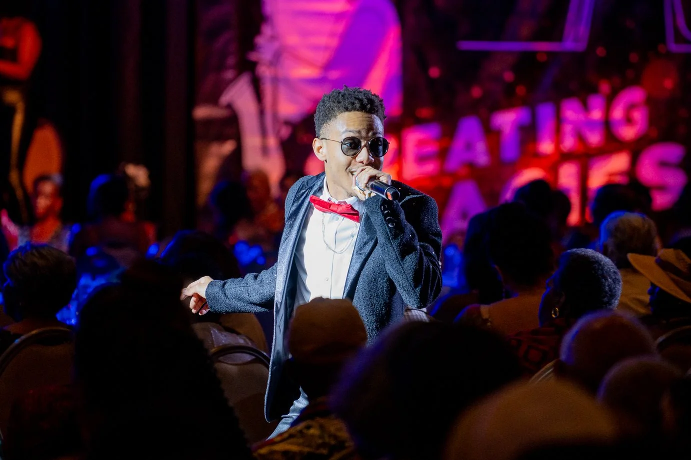 A man in a tuxedo with a red bow tie and sunglasses singing into a microphone at a comedy show, with an audience and vibrant stage background.