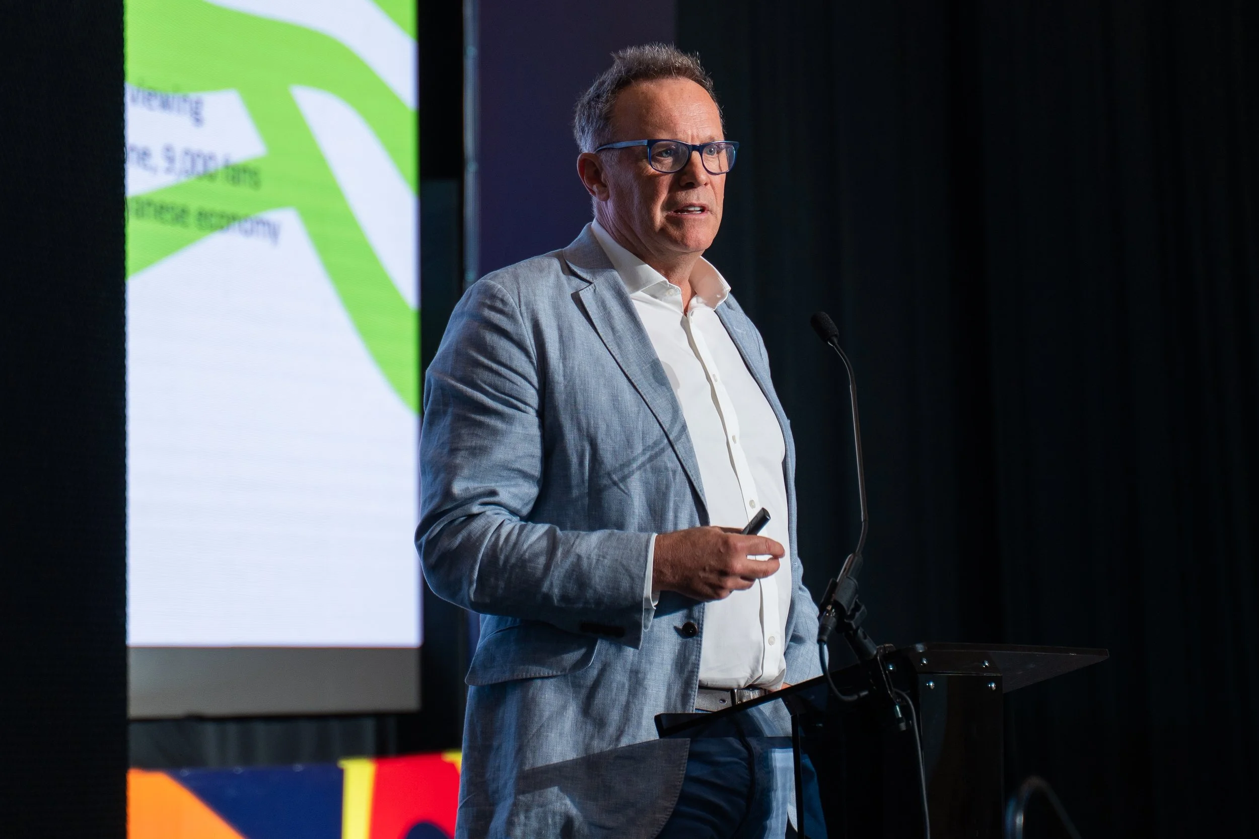 A man with glasses and a light gray blazer speaking at a presentation or conference, standing in front of a screen with colorful graphics.
