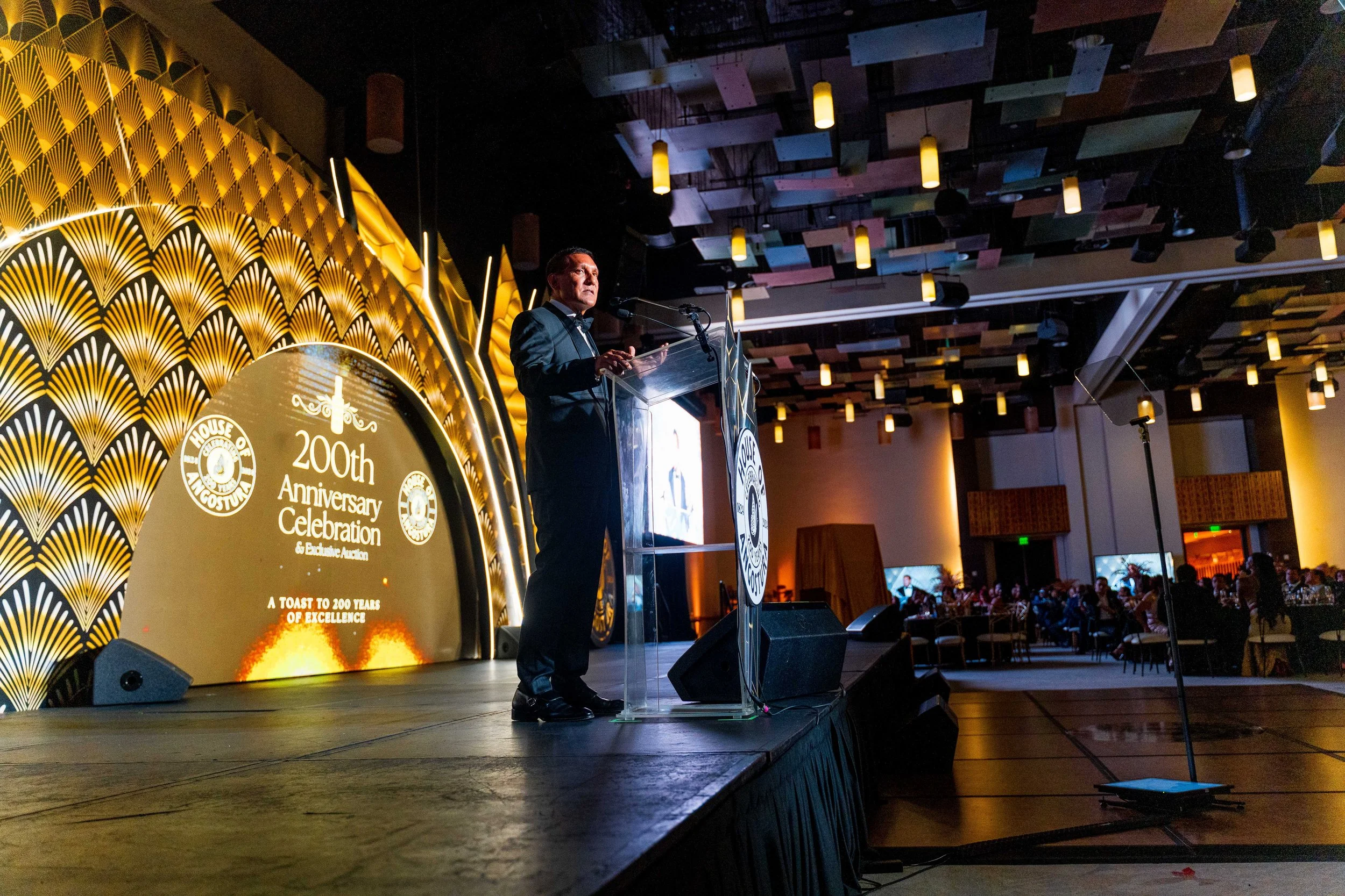 A man in formal attire giving a speech at an event with a stage decorated with a large emblem commemorating the 200th anniversary of the House of Tinuvelas, featuring golden fan patterns and text celebrating 200 years of excellence. The event is atte