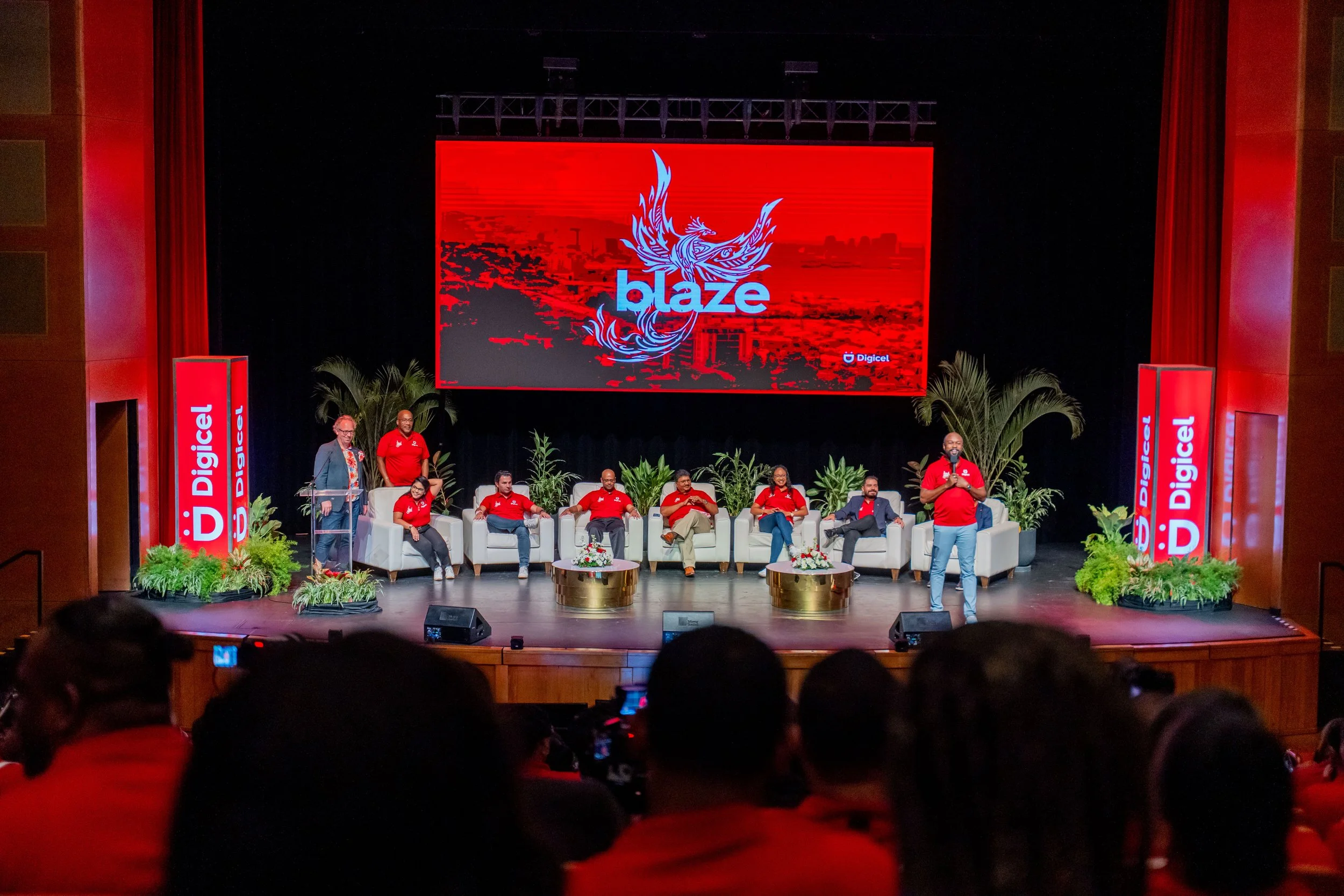 A stage with a large screen displaying the Blaze logo and a cityscape background. Several people are seated on white chairs, and one person is standing and speaking. The stage is decorated with plants and red Digicel banners on the sides.