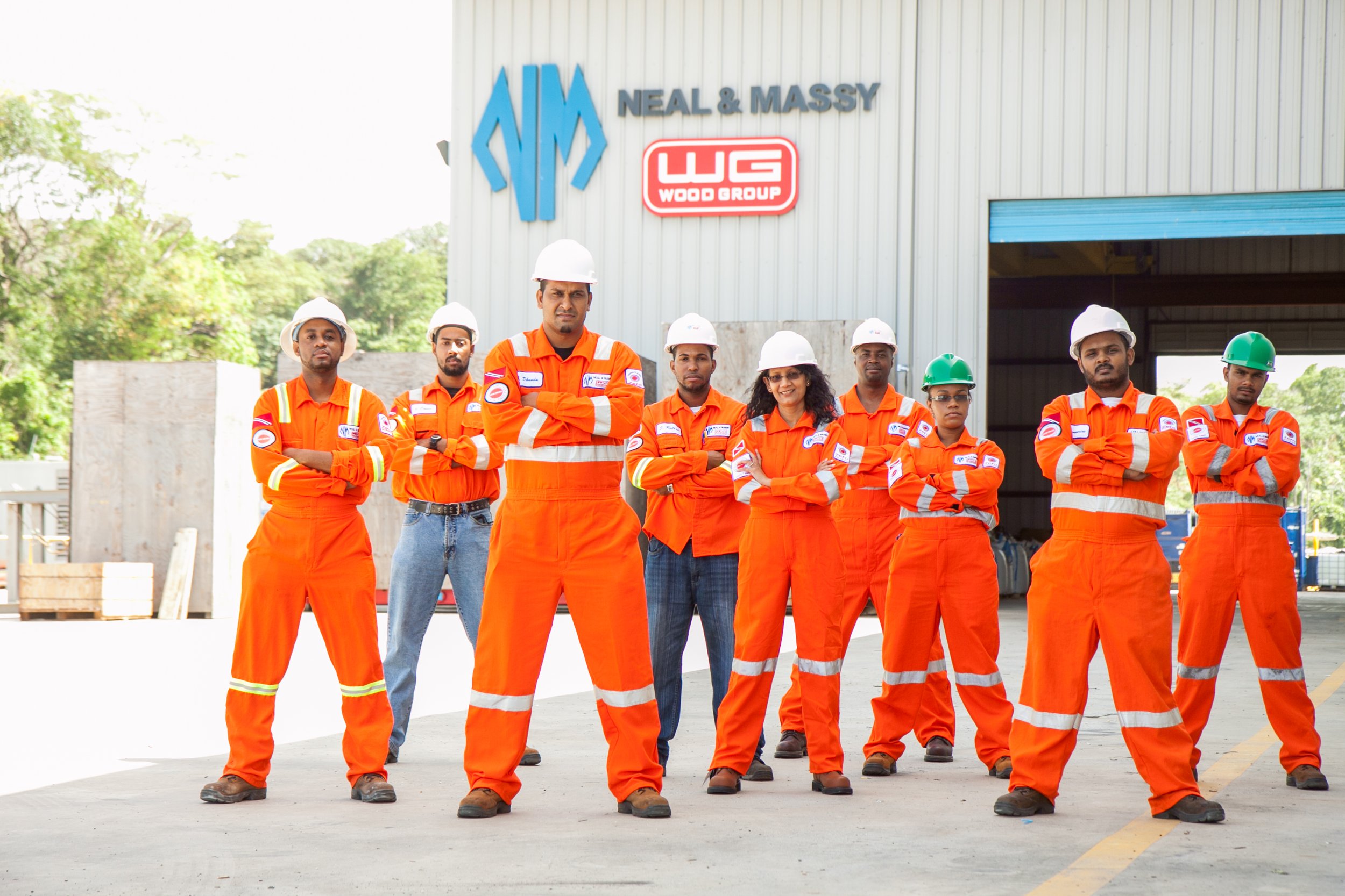 Group of construction workers standing with arms crossed in front of a building with a sign reading 'Neal & Massy' and a logo, some wearing white and green hard hats, all in orange safety uniforms.