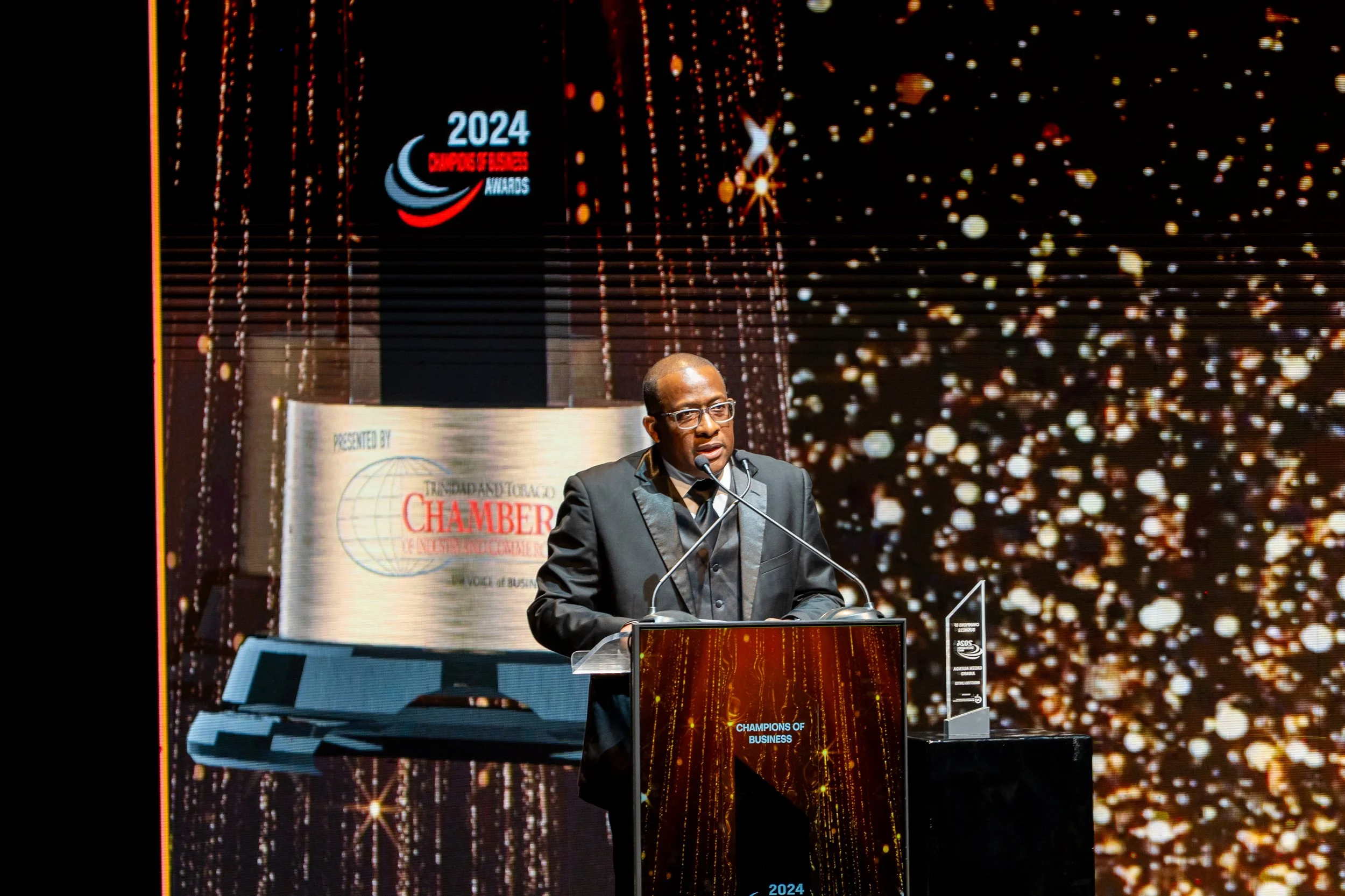 Man in black suit and glasses speaking at a podium during the 2024 Entrepreneurs of Excellence Awards, decorated with a large digital backdrop featuring gold and red sparkles, and logos including 'Chamber of Commerce' and 'Champions of Business.'.