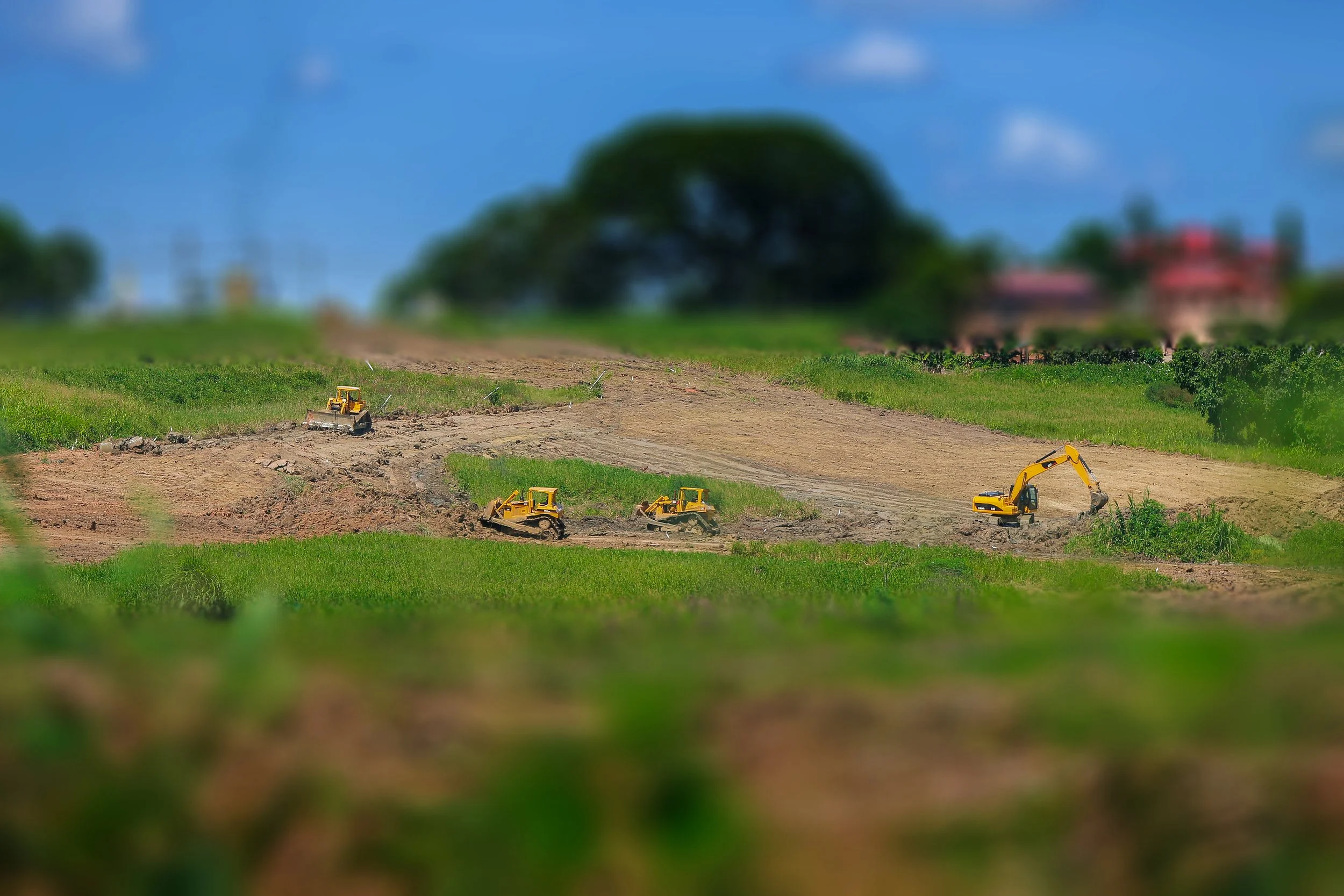 Construction site with yellow excavation equipment working on dirt field, surrounded by green grass, with trees and buildings in the background under blue sky.