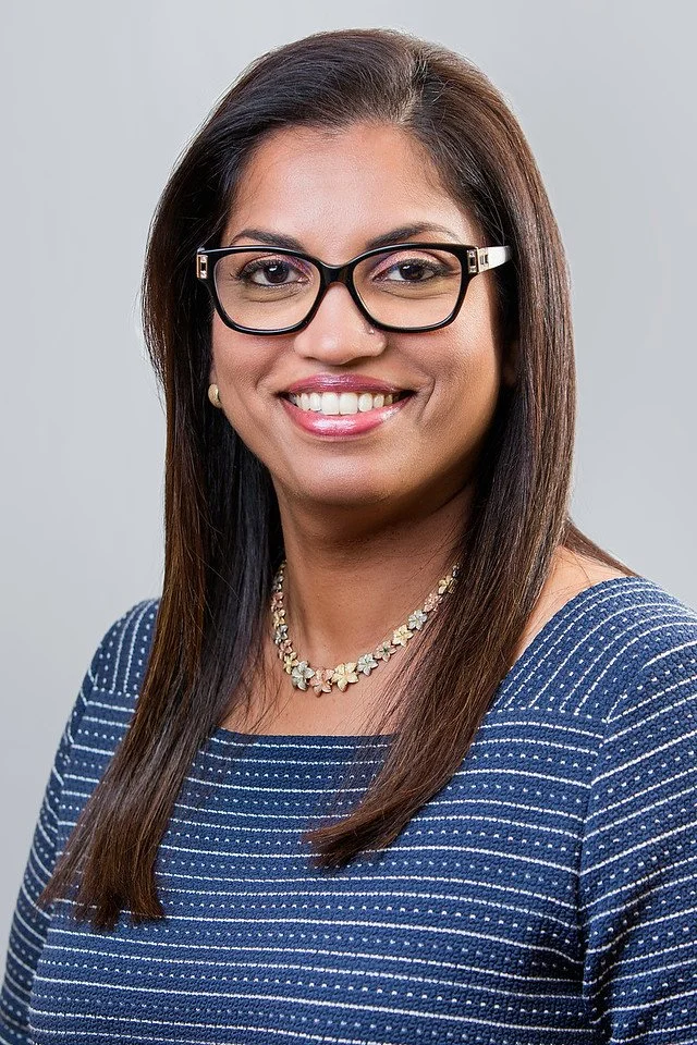 A woman with long brown hair, wearing black glasses, a blue and white striped top, and a white floral necklace, smiling against a plain gray background.