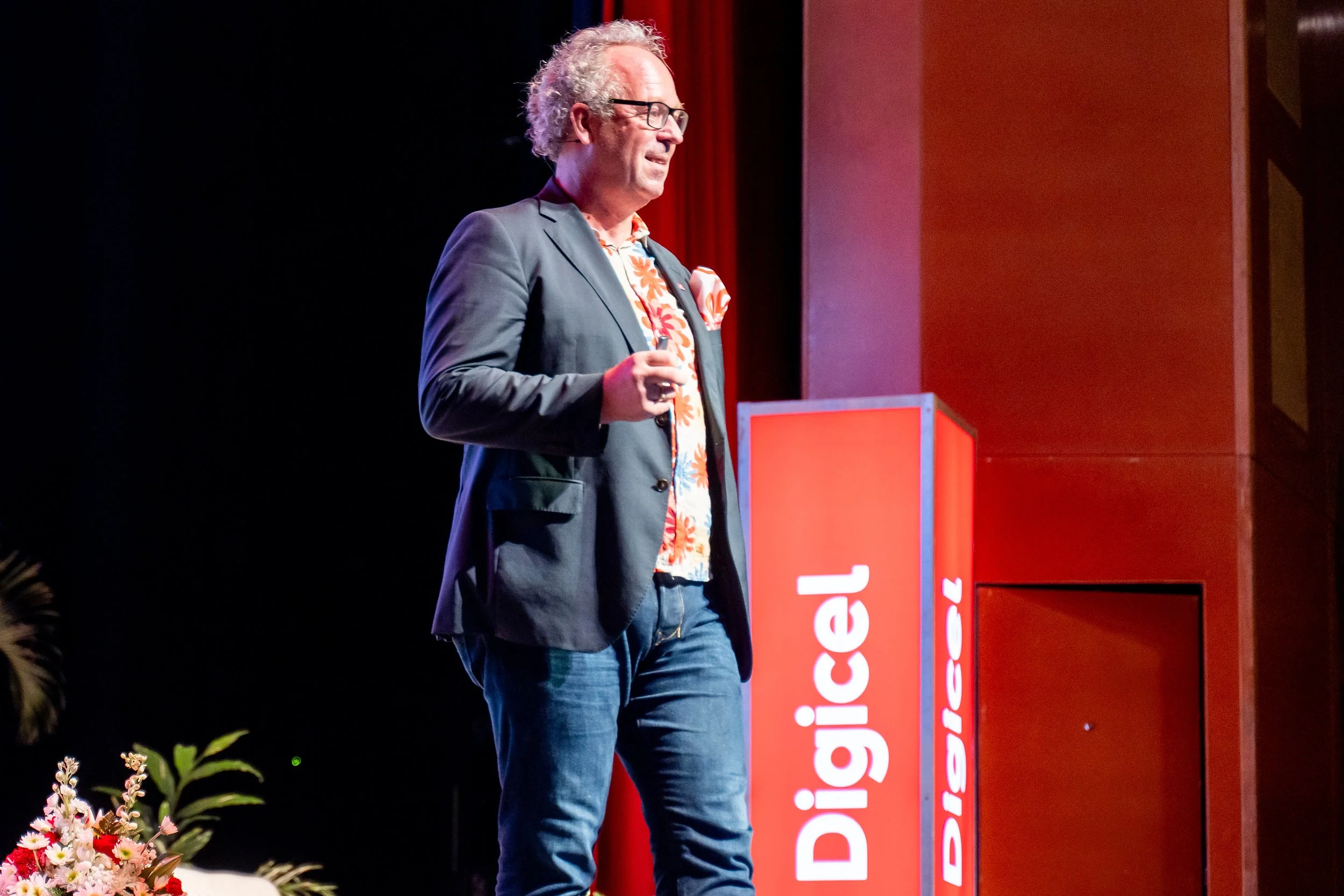 A man with curly gray hair, glasses, wearing a dark blazer and a floral shirt, speaking at a podium during an event, with a red banner and flowers in the foreground.