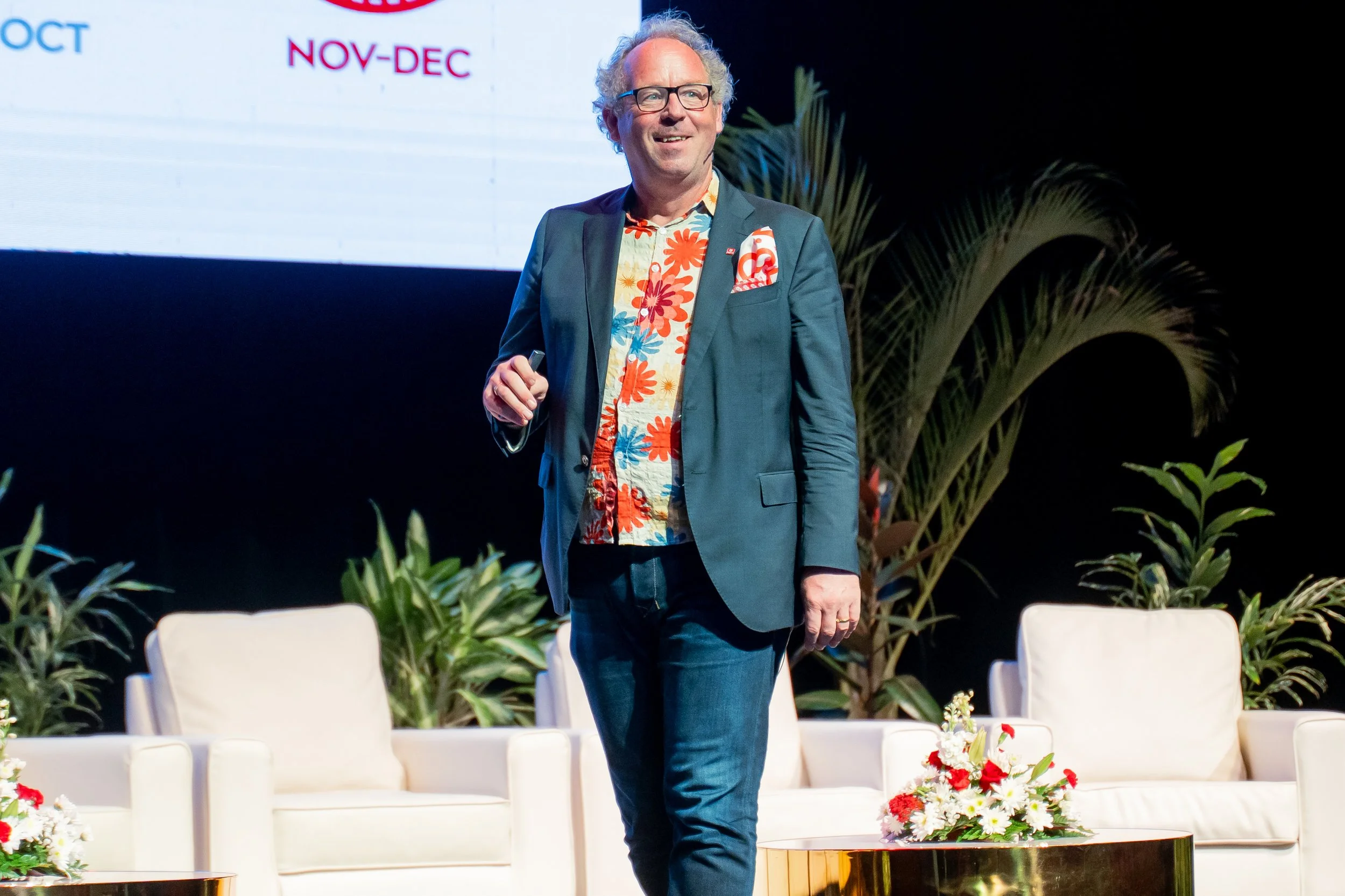 A man with curly gray hair and glasses smiling while standing on a stage with tropical plants, white chairs, and floral arrangements in front of him, with a screen behind displaying words 'OCT' and 'NOV-DEC'.