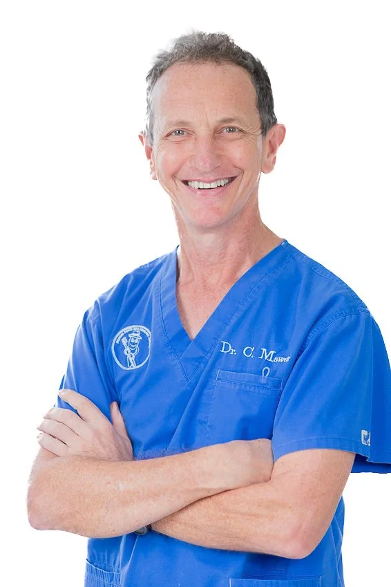 A smiling middle-aged man in blue medical scrubs with the words "Dr. C. Mauer" embroidered on it, standing with arms crossed against a plain white background.