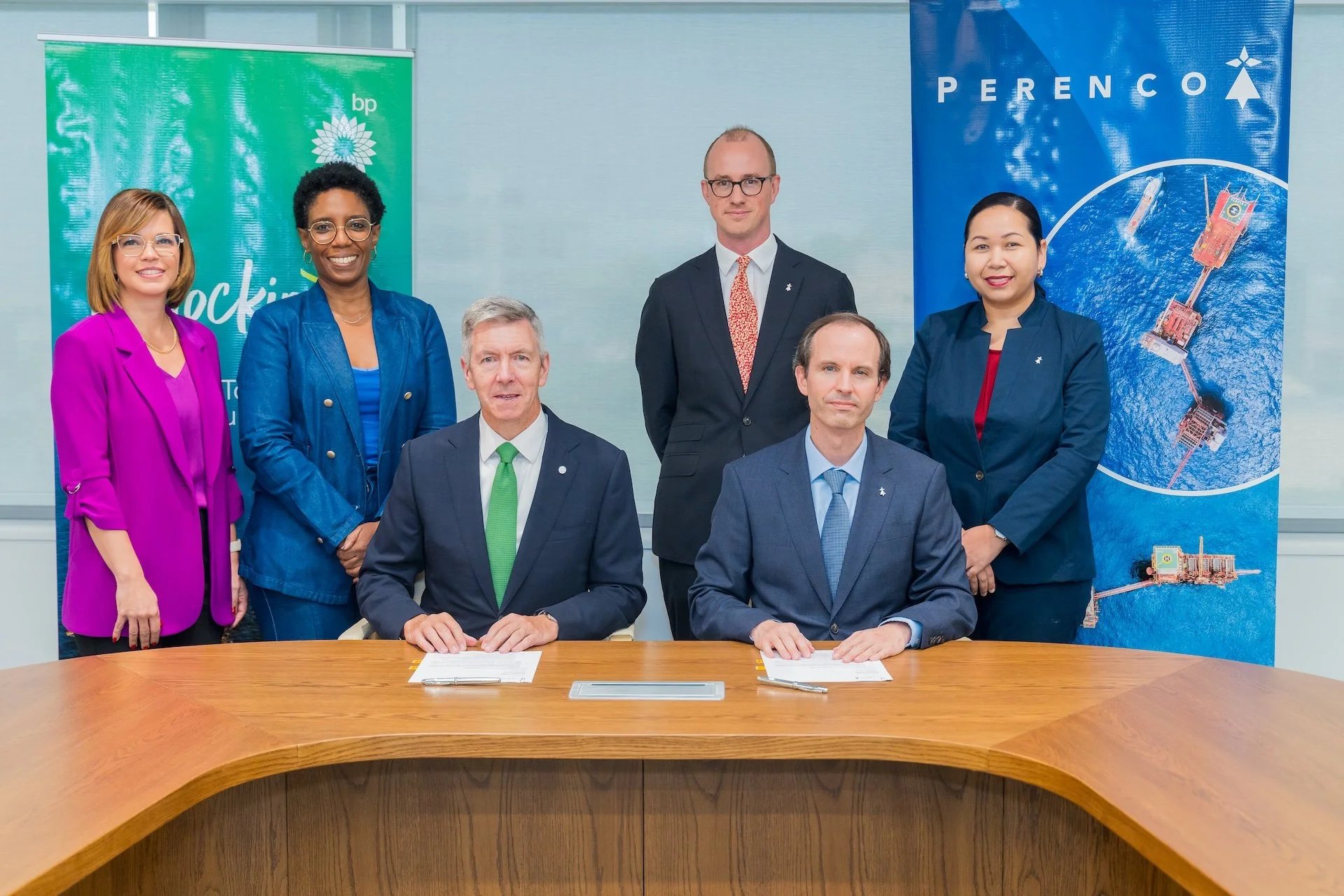 Seven diverse professionals are gathered in a conference room, with three seated at a wooden table and four standing behind them. Behind them are banners with corporate logos and a blue background with a satellite image. The seated individuals are me