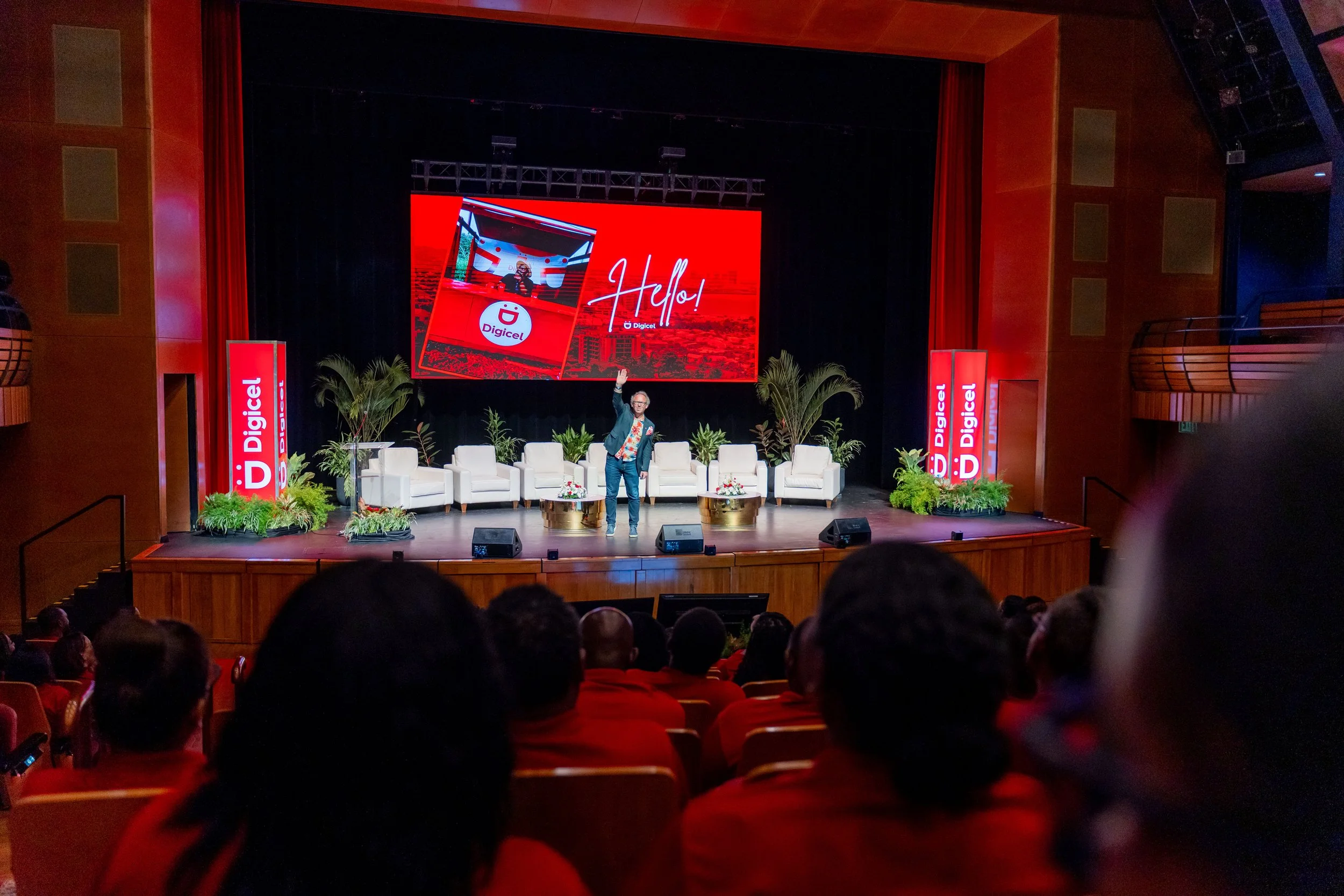A stage with a large screen displaying the Digicel logo and the word 'Hello!'. There are several chairs lined up on the stage, decorated with plants and flowers. A man is standing on the stage, waving to the audience.