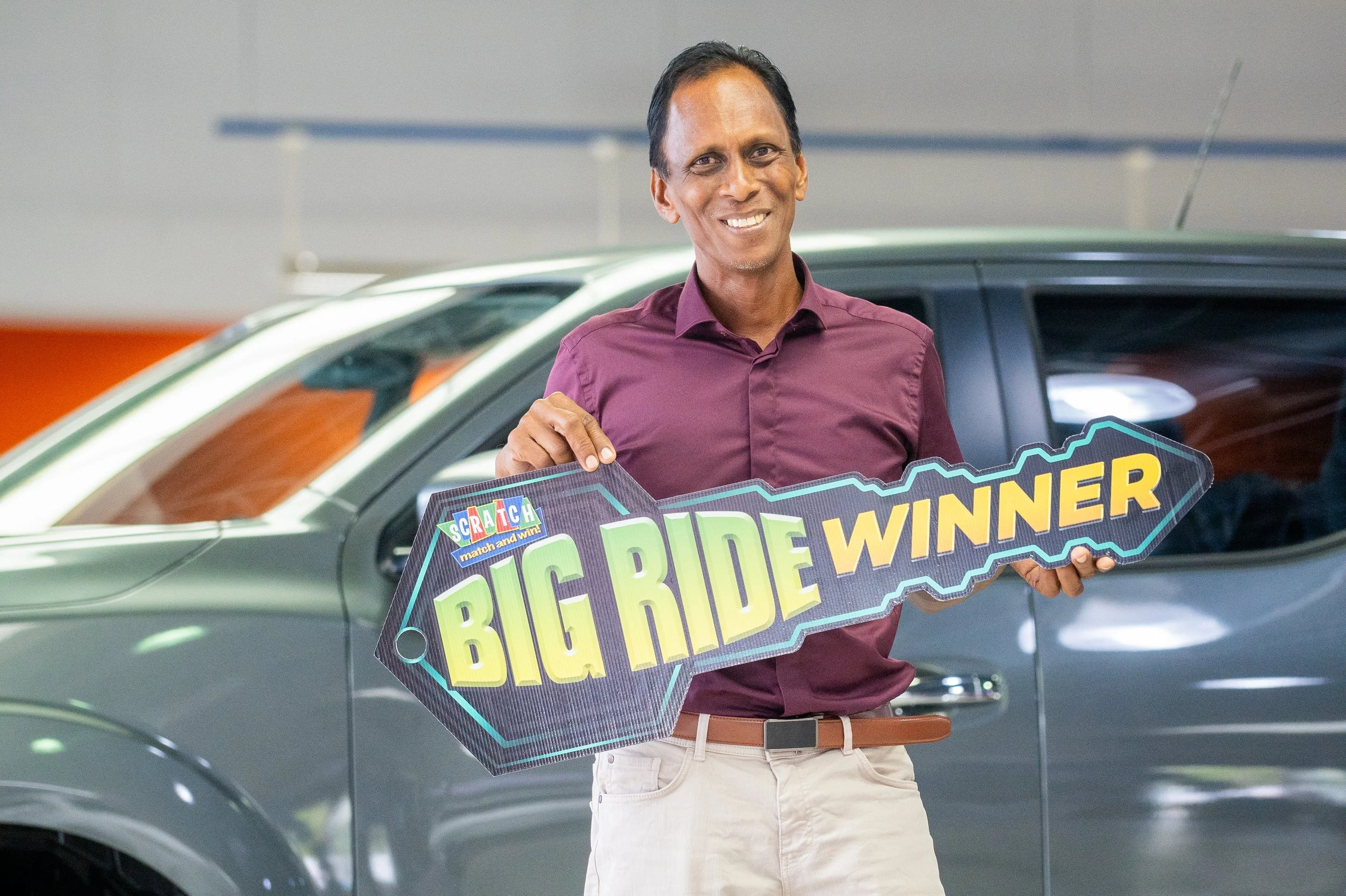 A man in a maroon shirt holding a 'Big Ride Winner' sign in front of a silver vehicle, smiling in an indoor setting.