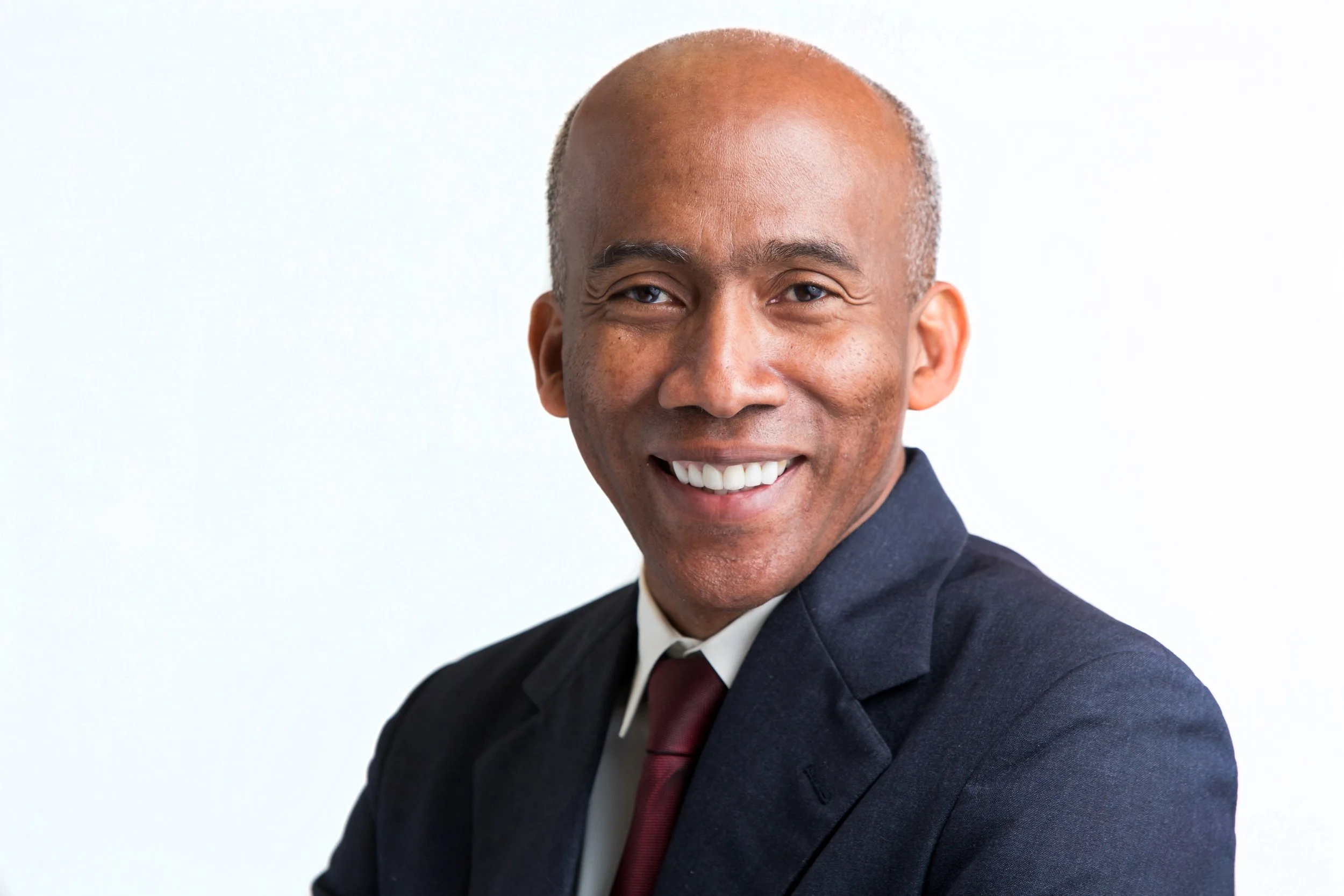 A smiling middle-aged man with a shaved head, wearing a dark suit, white shirt, and maroon tie, against a plain white background.