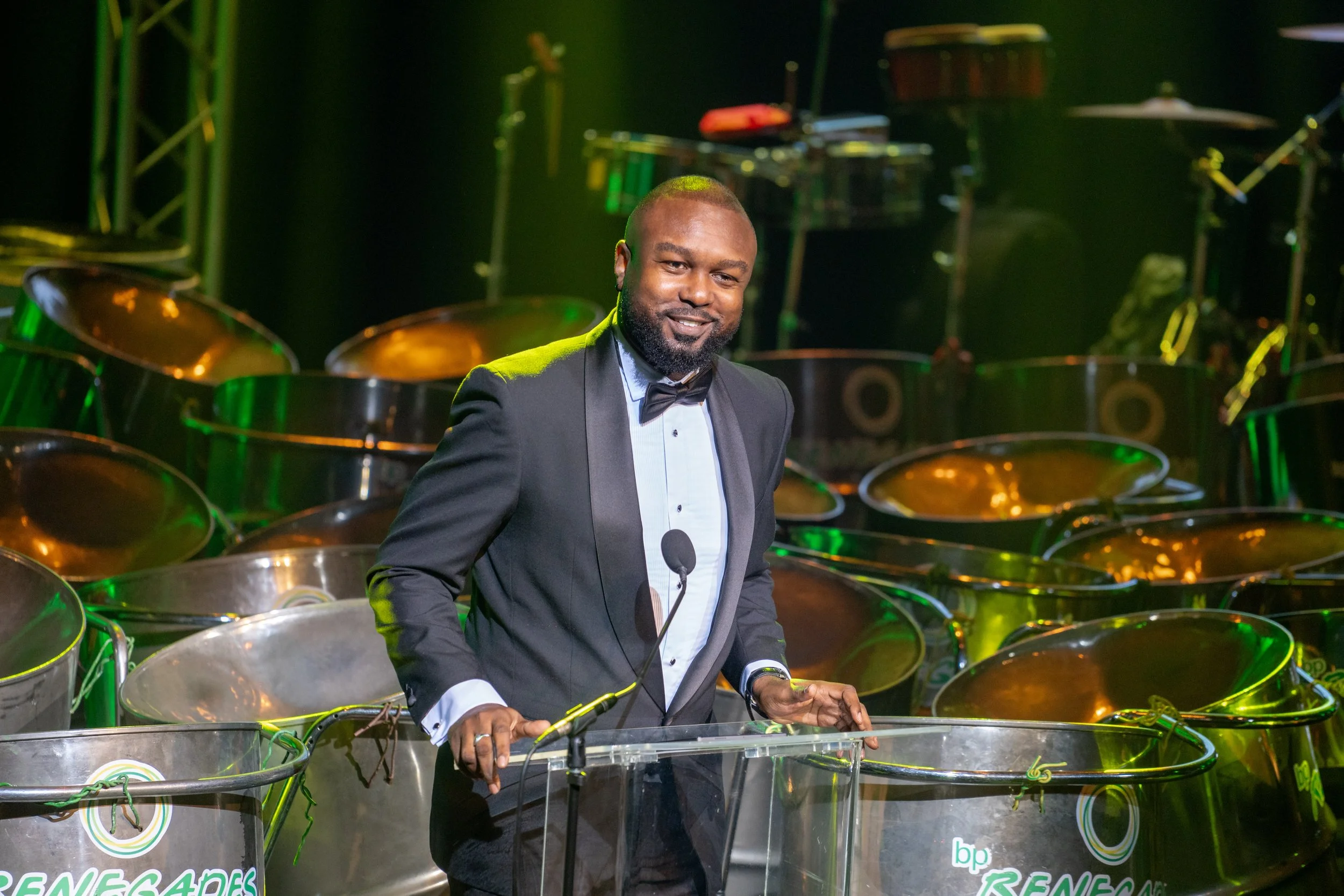 A man in a tuxedo playing steel drums on stage surrounded by multiple steel drums and musical instruments, with stage lighting.