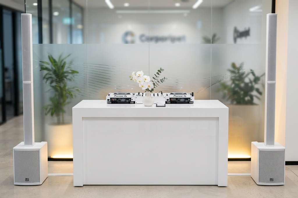 DJ equipment with a vase of white flowers on a white table, in front of a frosted glass wall with blurred logo or text, flanked by large white speakers in an indoor space.