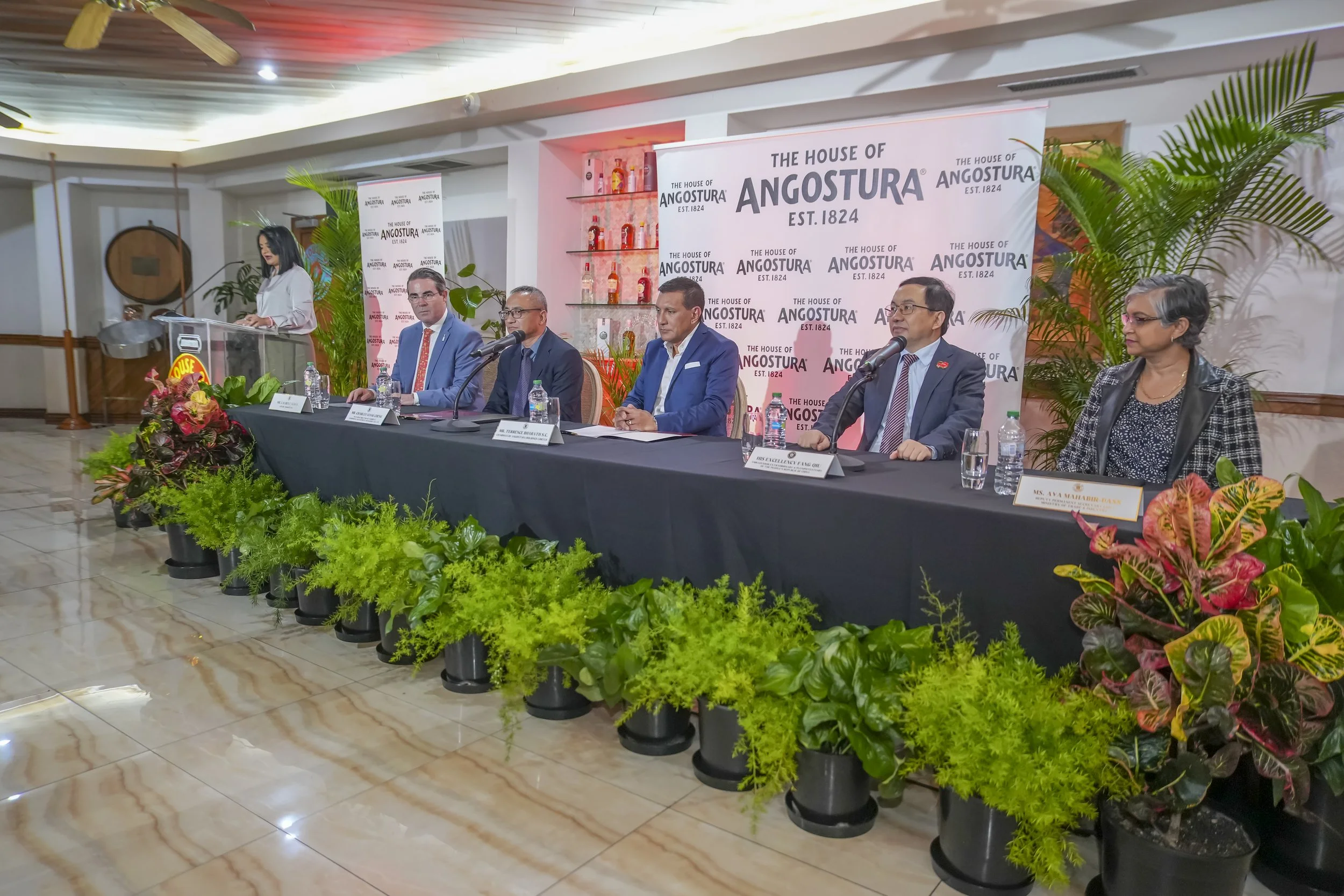 A formal event room with a panel of five people sitting at a table with microphones and water bottles, and a woman speaking at a podium. The backdrop displays the logo of The House of Angostura, established in 1824, with shelves of bottles and greene