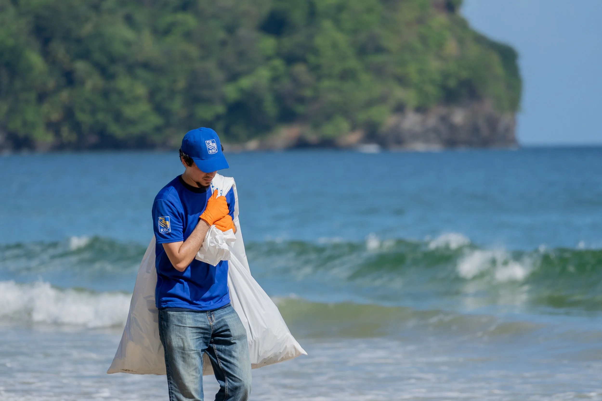 A person in a blue cap and shirt, wearing orange gloves, walks along the beach carrying a large white trash bag, with green hillside and ocean in the background.