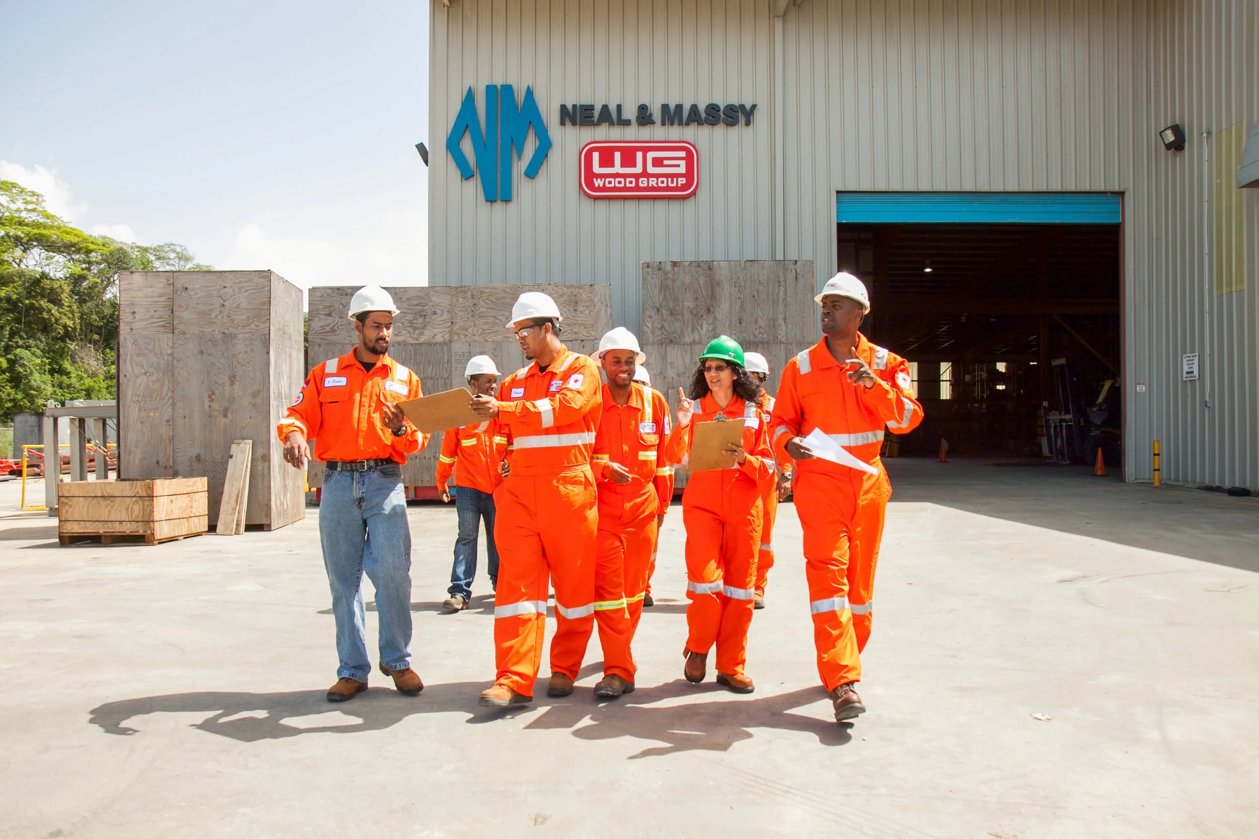 Group of construction workers walking outside of a building with a sign that reads 'NEAL & MASSY' and 'WOOD GROUP.' The workers are wearing orange safety uniforms and white hard hats, some holding blueprints and clipboards.