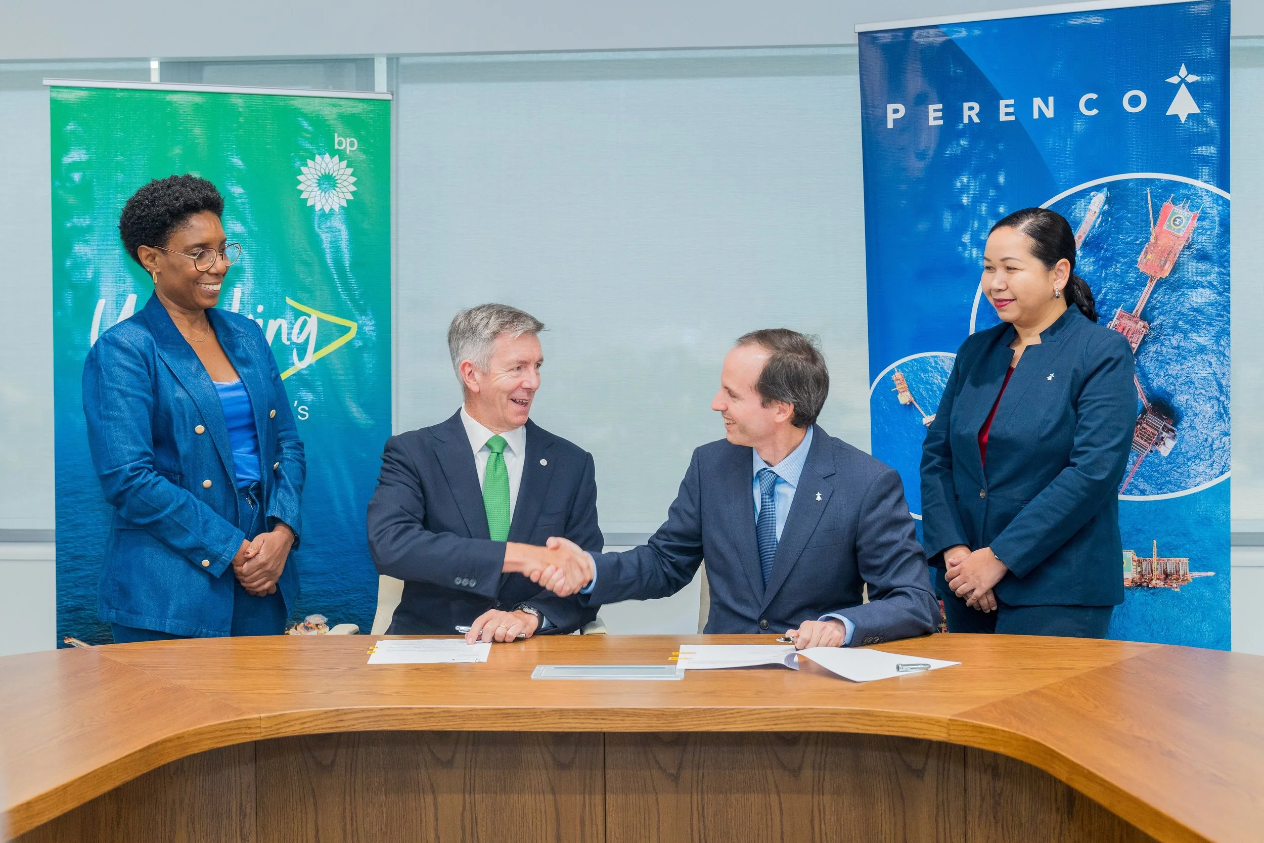Two men and two women in business attire at a signing event, with handshake between the men, in a conference room with banners for BP and Perenco in the background.