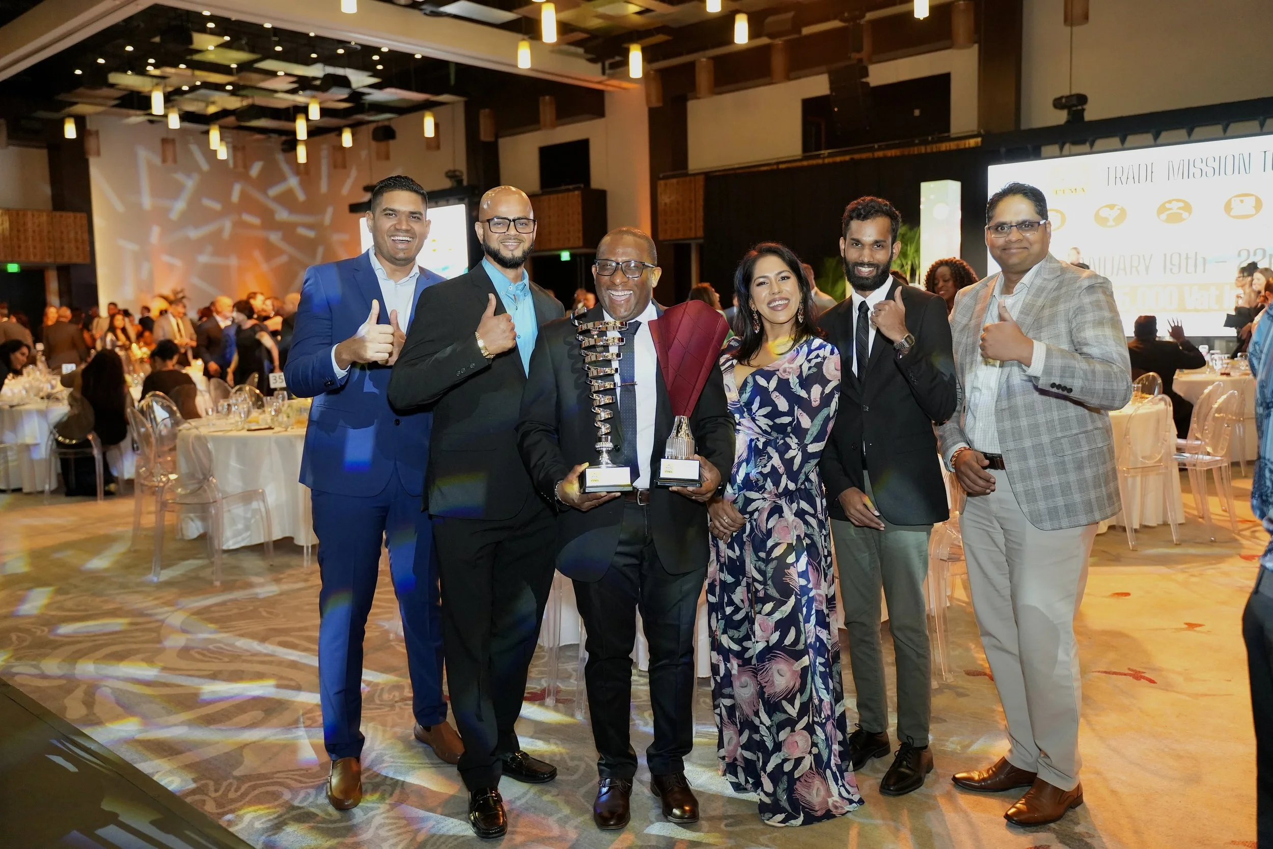 Group of six diverse people at an award ceremony, with three men dressed in suits and a woman in a floral dress, holding trophies and smiling in a banquet hall with tables and a stage in the background.