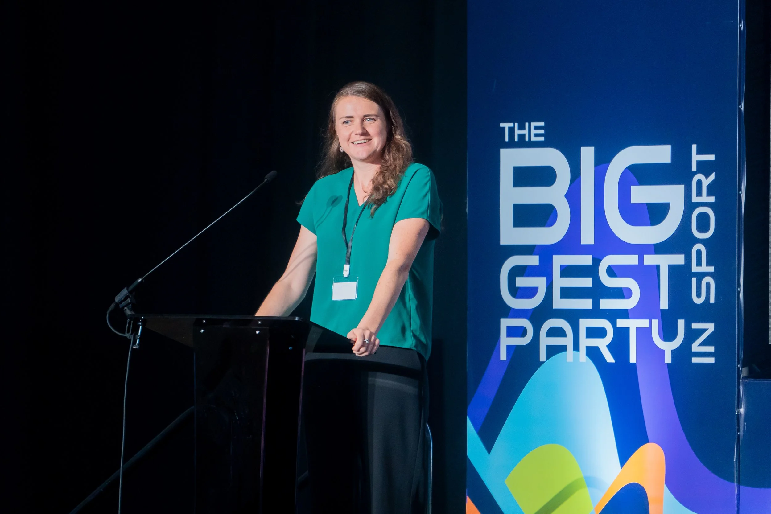 A woman standing at a podium with a microphone, smiling, during a conference or event, with a colorful logo backdrop that says "The Biggest Party" and other partially visible words.