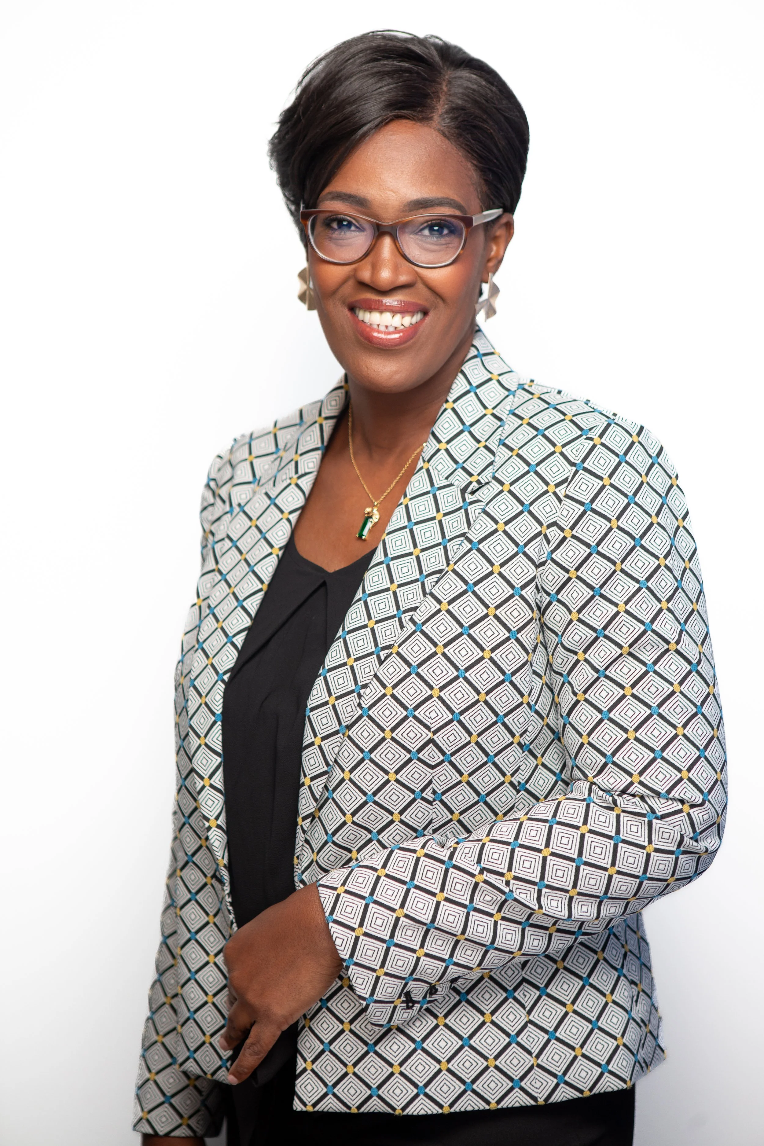 Portrait of an African American woman with short hair, wearing glasses, a black top, and a patterned blazer, smiling against a white background.