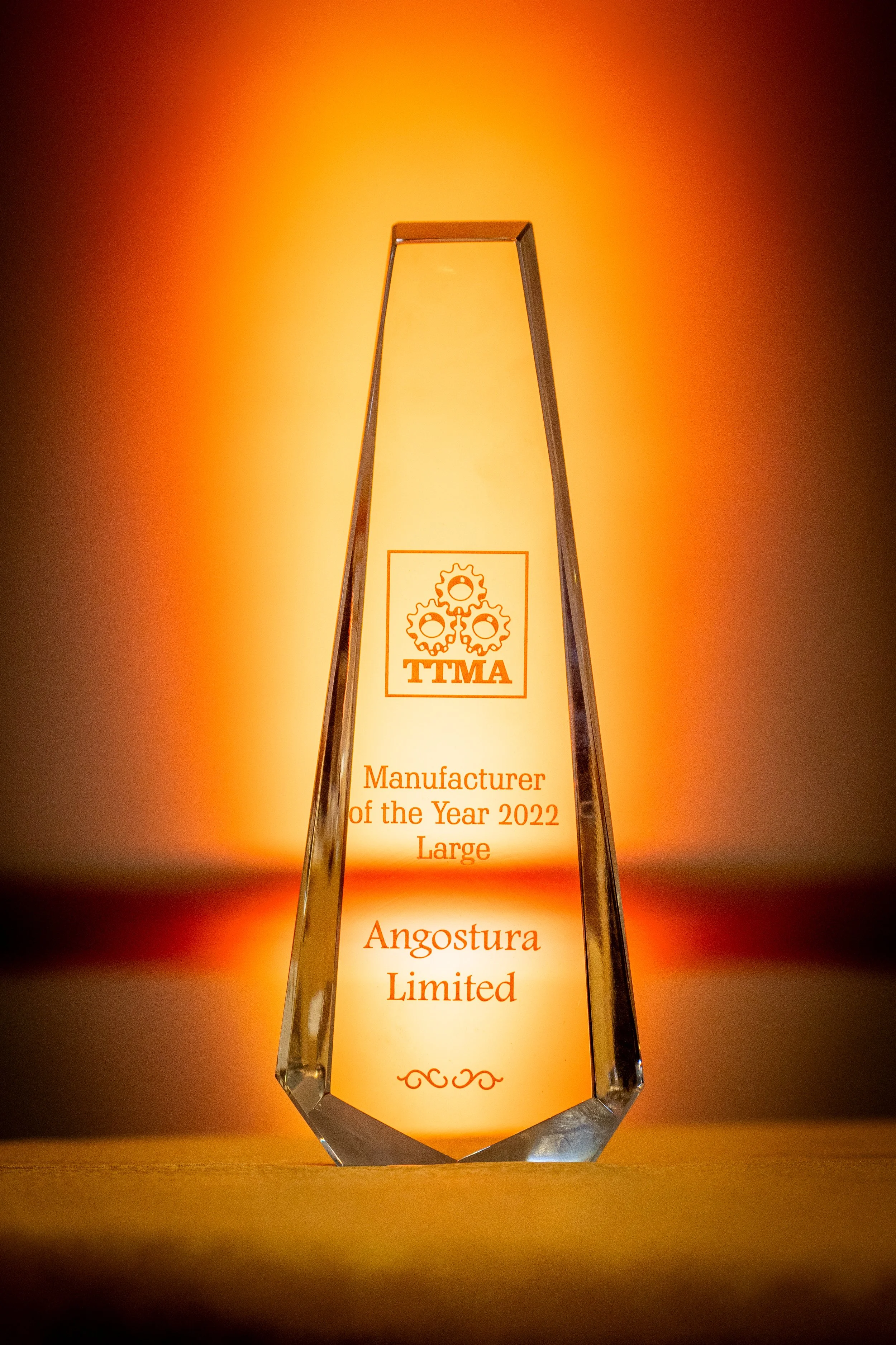 A clear, tall award trophy with engraved text and logo, illuminated from behind, standing on a wooden surface against a warm orange background.