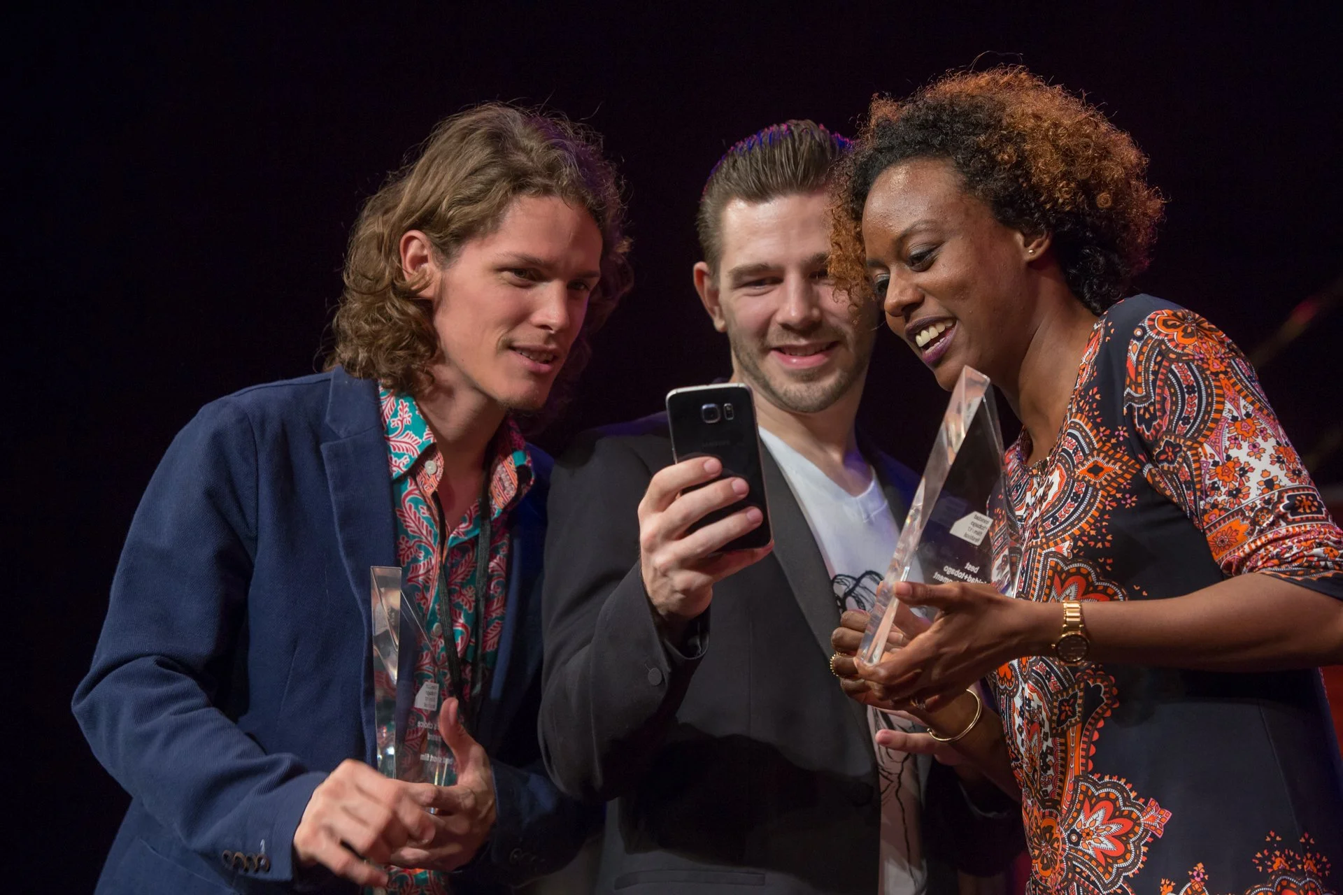 Three people on stage looking at awards and a phone, celebrating their achievements.