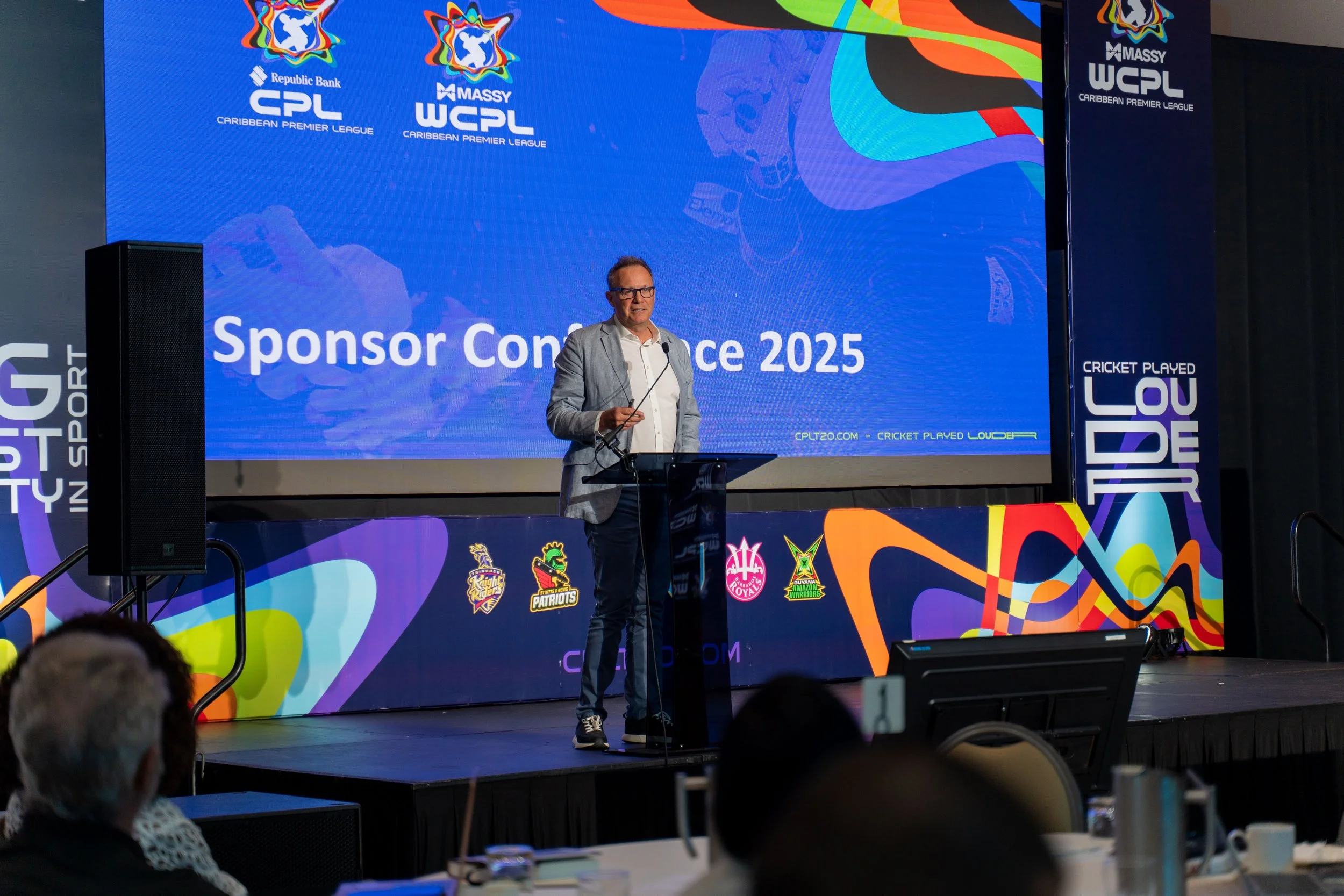 A man in a light gray blazer and dark pants is speaking at a conference on a stage with a large colorful screen behind him that displays the words 'Sponsor Conference 2025' and logos of cricket teams and the Caribbean Premier League (CPL). The stage 