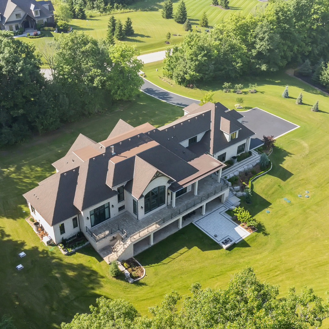 Aerial view of a large modern house with a landscaped backyard, patio, and surrounding trees in a suburban neighborhood.