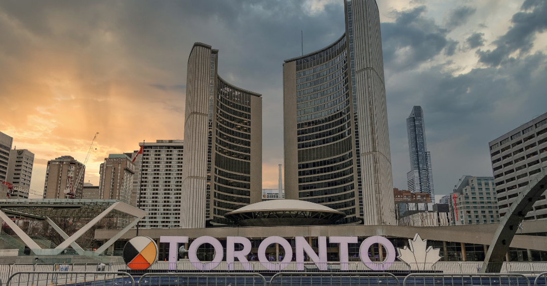 Downtown Toronto skyline with modern high-rise buildings, a large sign spelling 'TORONTO' with a maple leaf symbol, and a sunset sky.
