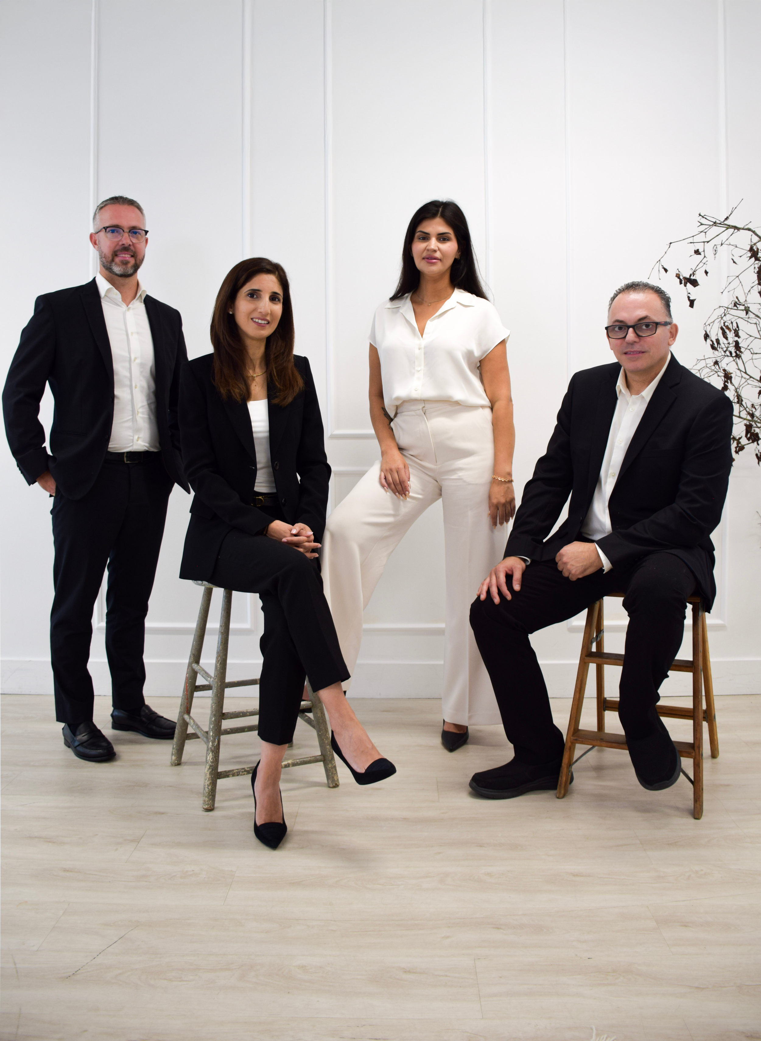 Group of five professionals, two men and three women, in formal business attire, posing in a modern, minimalist office with white walls and wooden flooring.